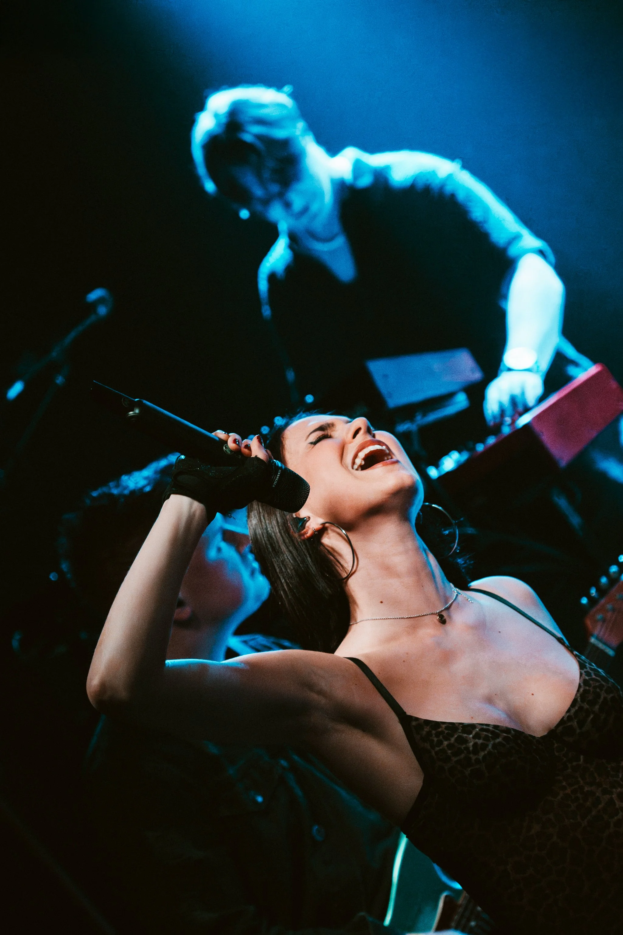 A woman singing into a microphone on stage, with a bright expression, wearing a spaghetti strap top and jewelry. A man in the background appears to be near a keyboard or electronic device, surrounded by dim stage lighting.
