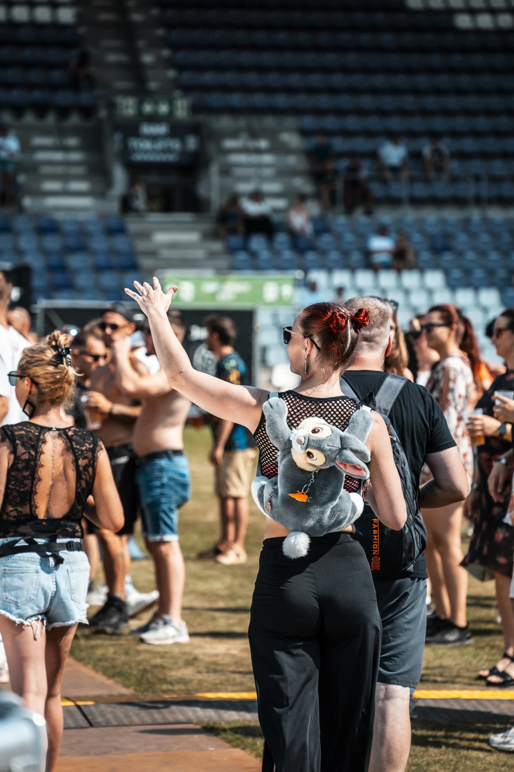 A diverse group of people standing and talking outdoors at a music festival or concert, with some sitting in the background on stadium seating.