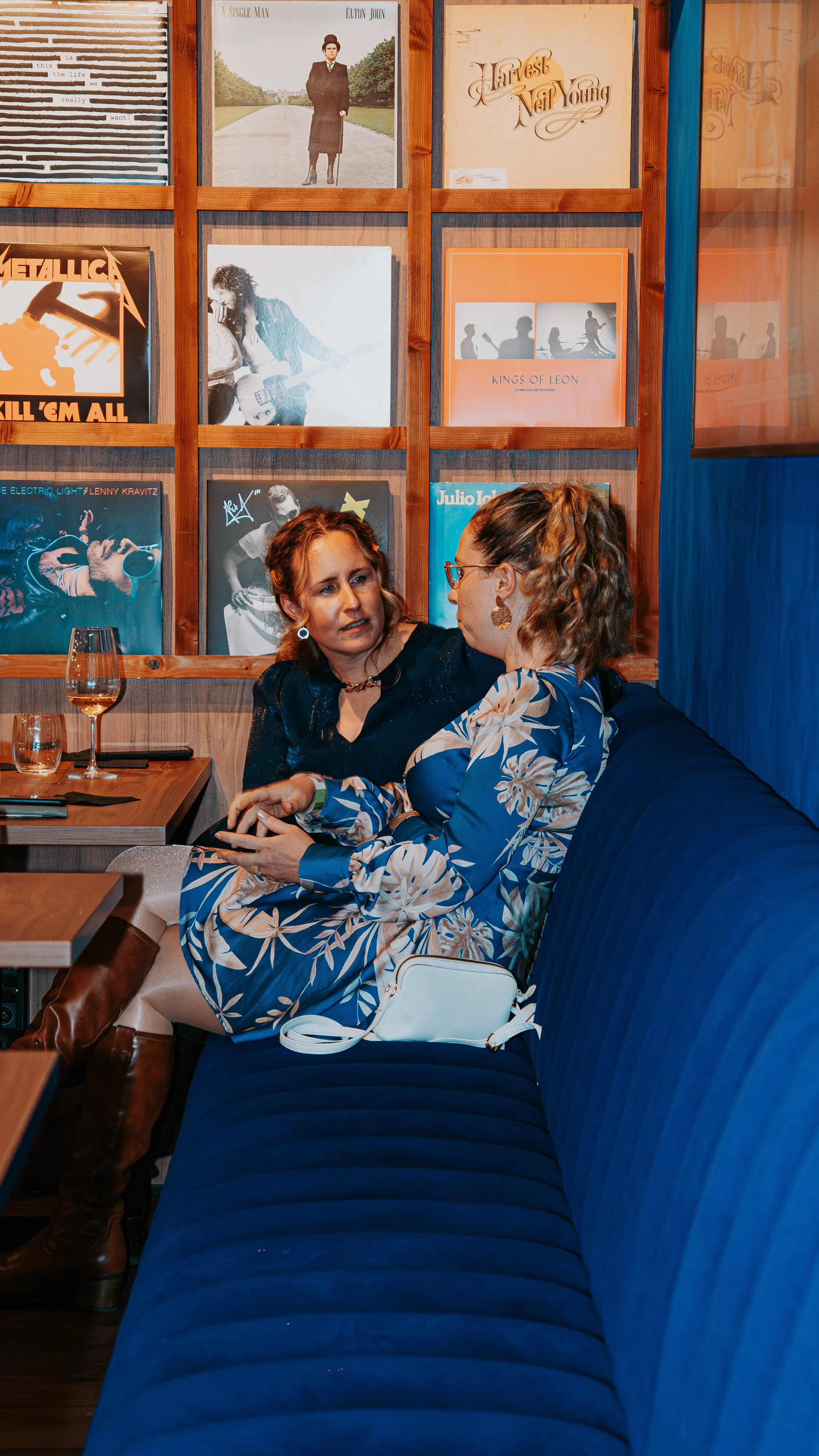 Two women are sitting in a restaurant or bar, engaged in conversation. One woman has curly hair, wears a dark top, and has earrings. The other woman has glasses, wavy hair pulled back, and is wearing a floral blouse. There is a table with a wine glas