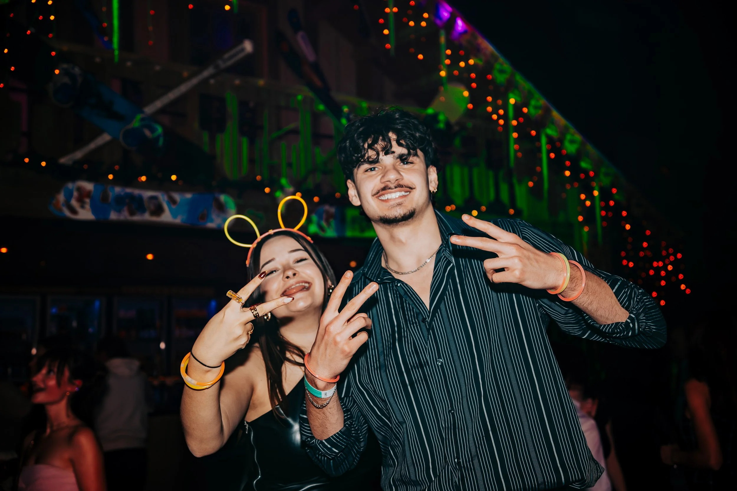 A young man and woman at a party or nightclub, smiling and posing for the camera with their hands making peace signs. The background is decorated with colorful lights and people are dancing.