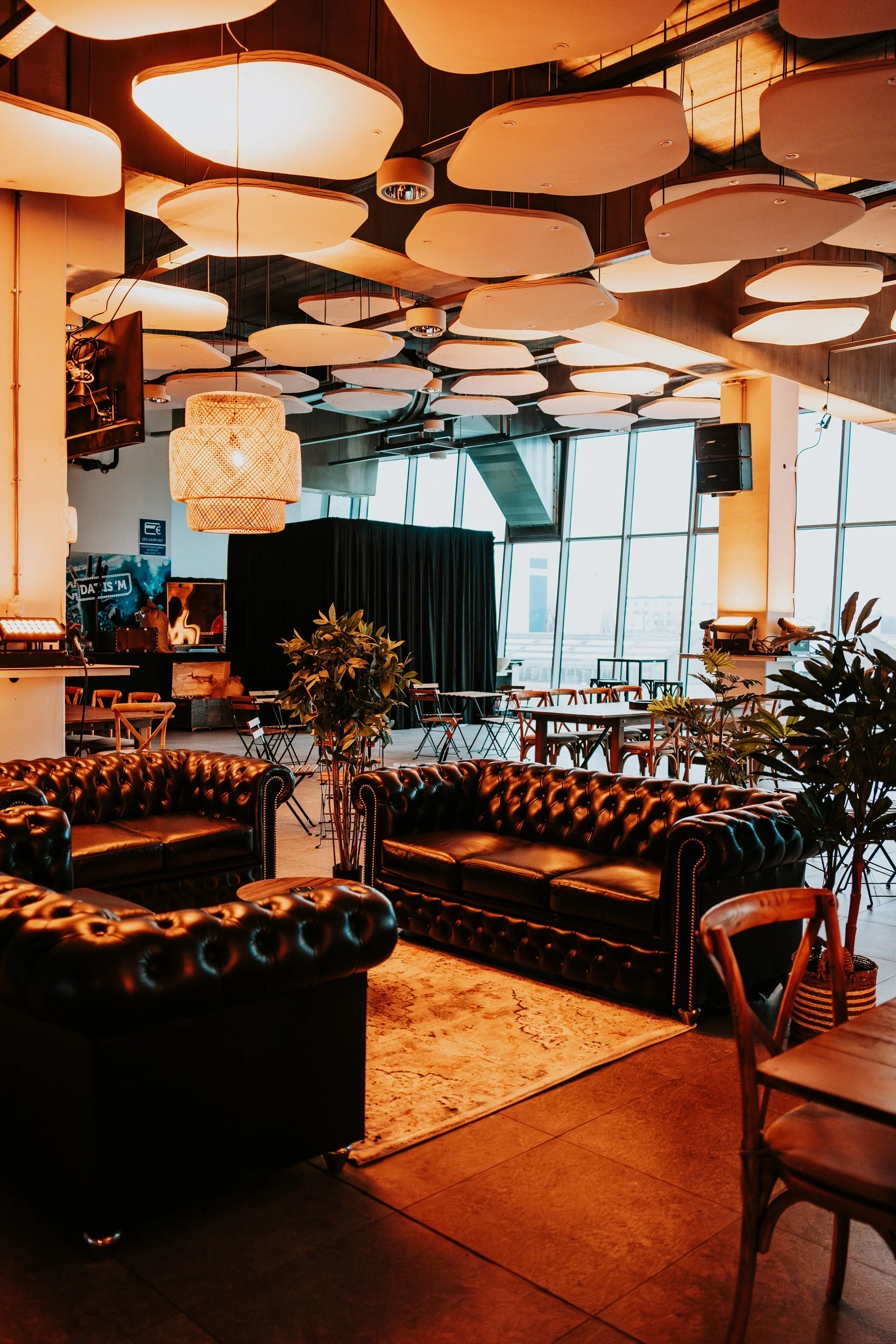 An interior view of a modern, stylish lounge or bar area with large, warm-colored ceiling lights, black leather tufted sofas, wooden chairs, plants, and large floor-to-ceiling windows.