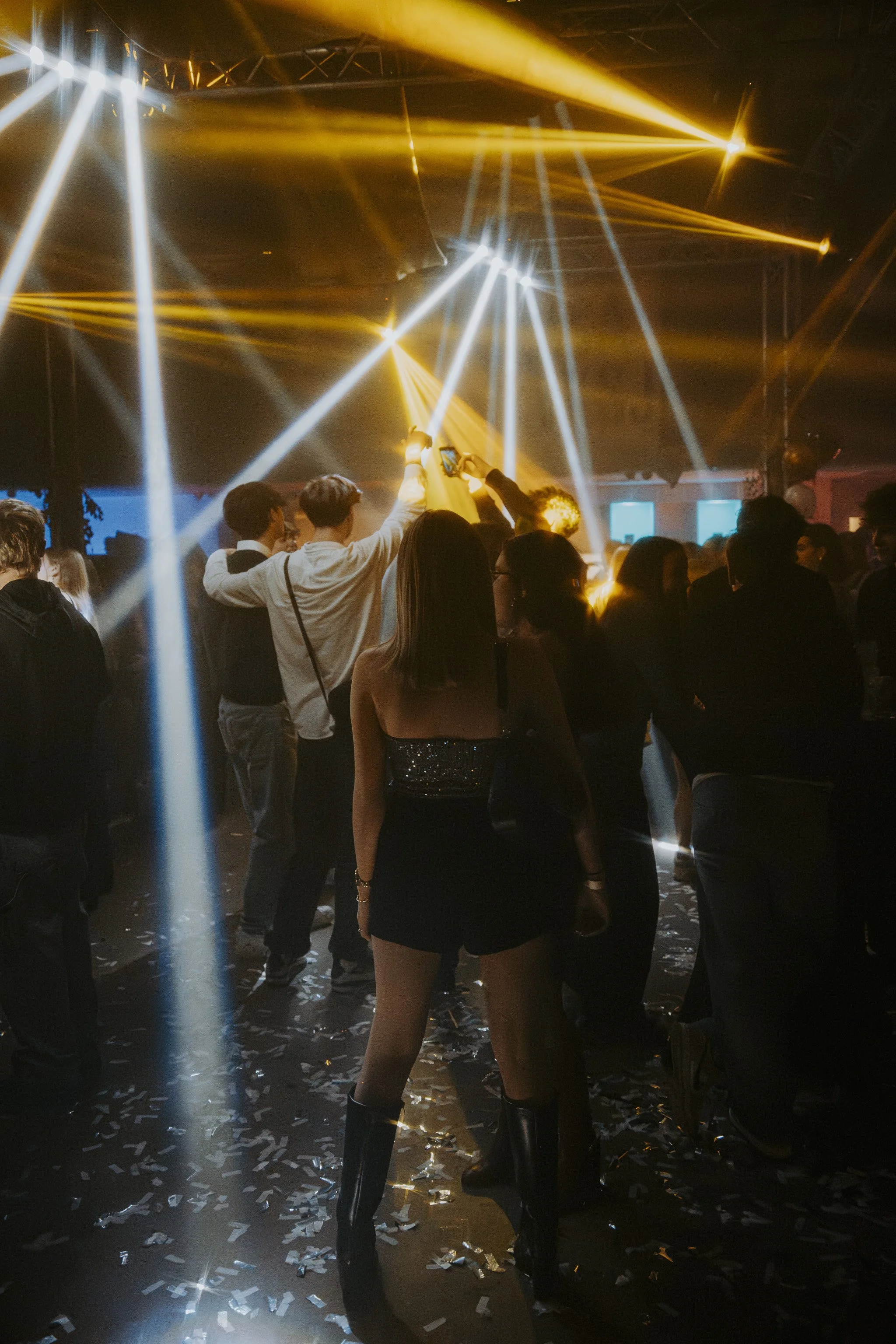 People dancing and taking photos at a party with yellow and white stage lights, confetti on the floor, and a dark background.