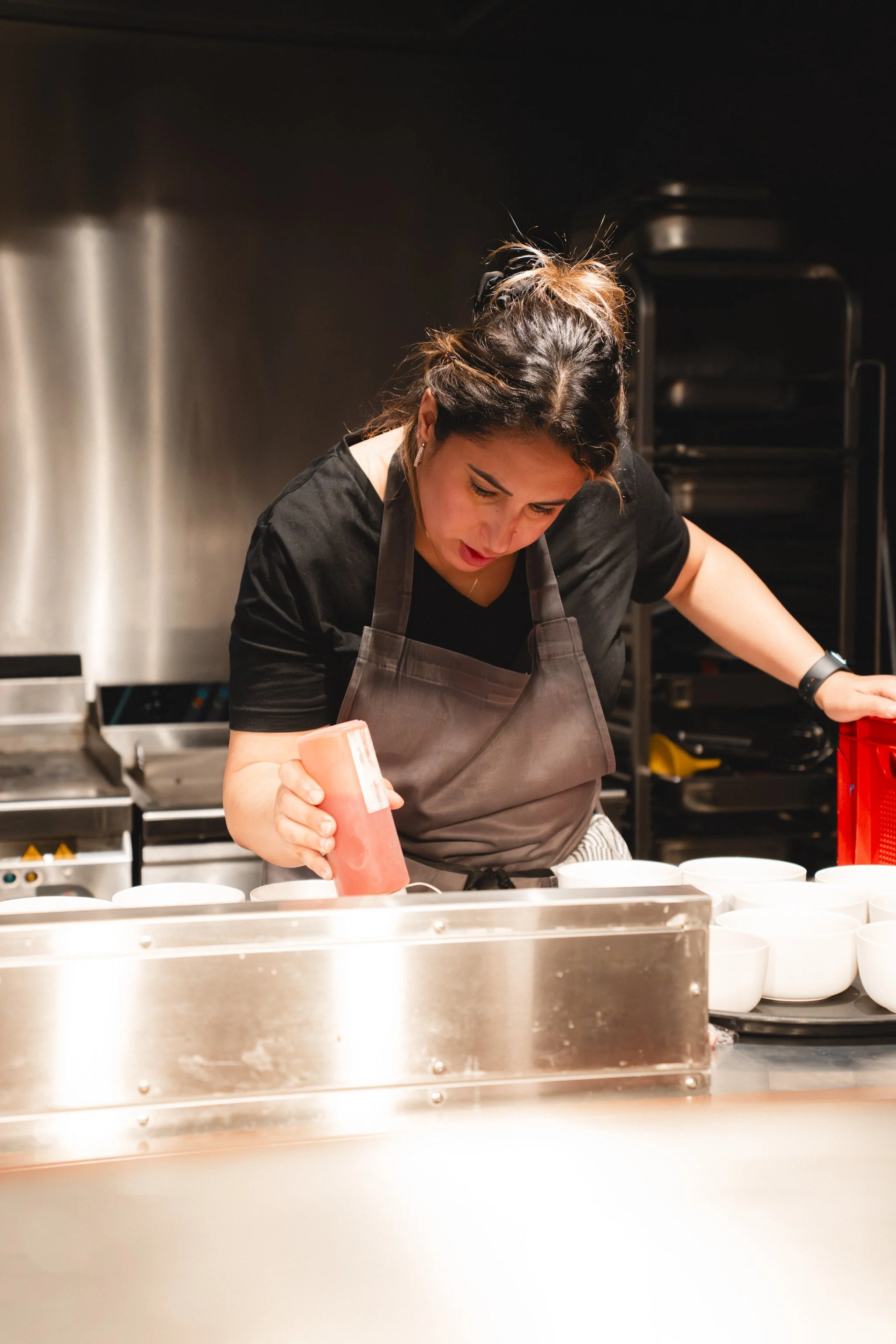 A woman wearing a black shirt and gray apron is in a kitchen, squeezing a pink substance from a bottle into a white bowl, with several white bowls in front of her.