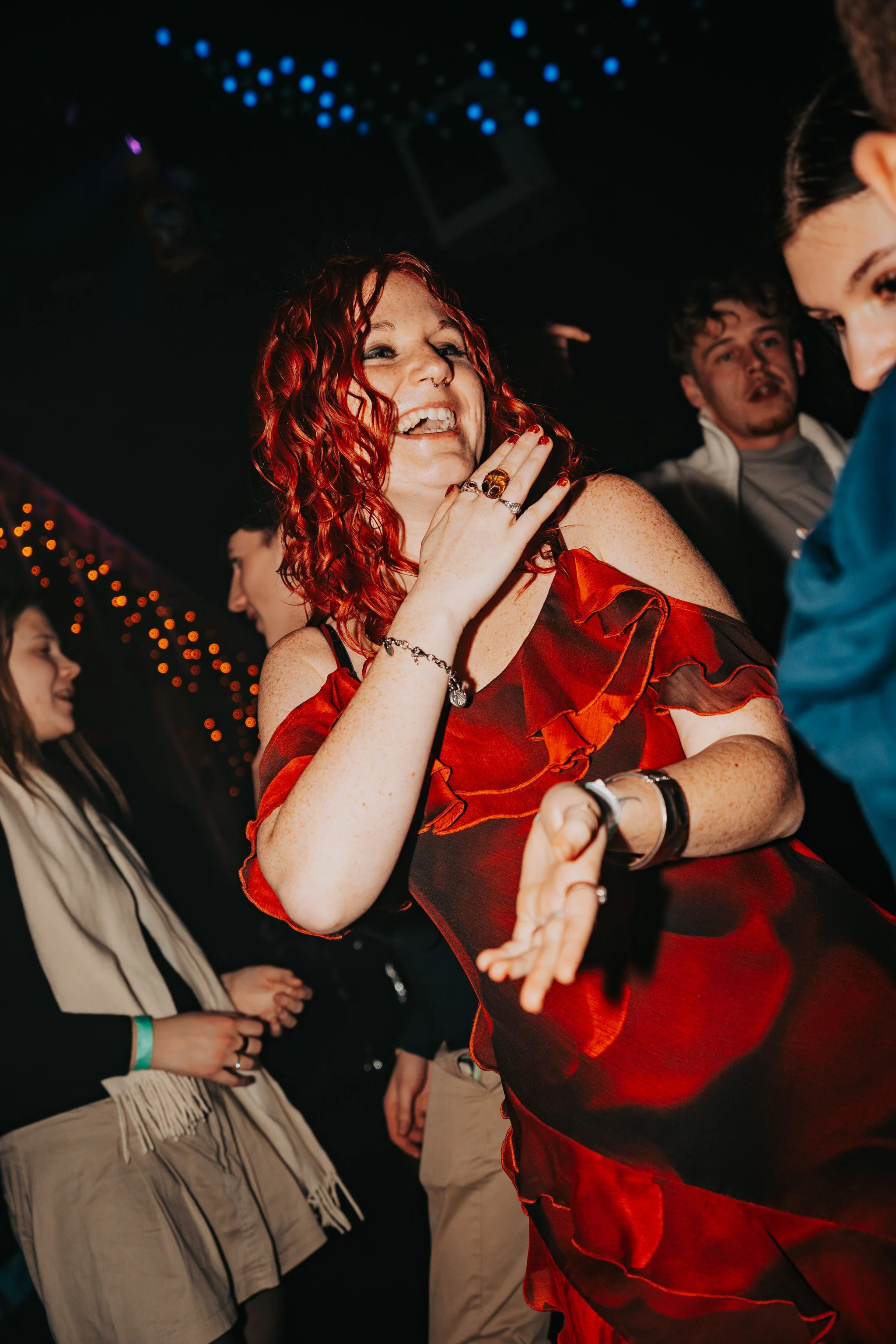 Group of people at a party or club, with one woman with red hair and a tattoo, smiling and touching her neck, wearing a red dress with ruffle details and assorted jewelry, in a setting with dark lighting and colorful lights.