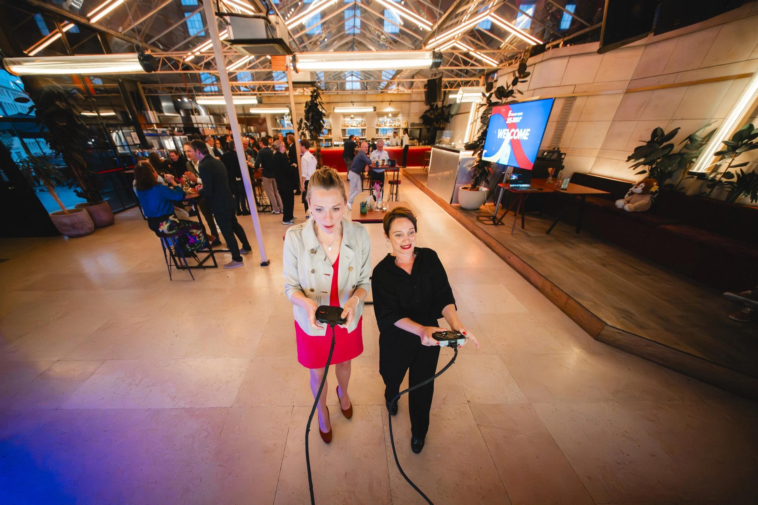 Two women are walking in a lively indoor space, holding gaming controllers. The environment features modern decor, plants, and bright lighting, with other people socializing in the background.