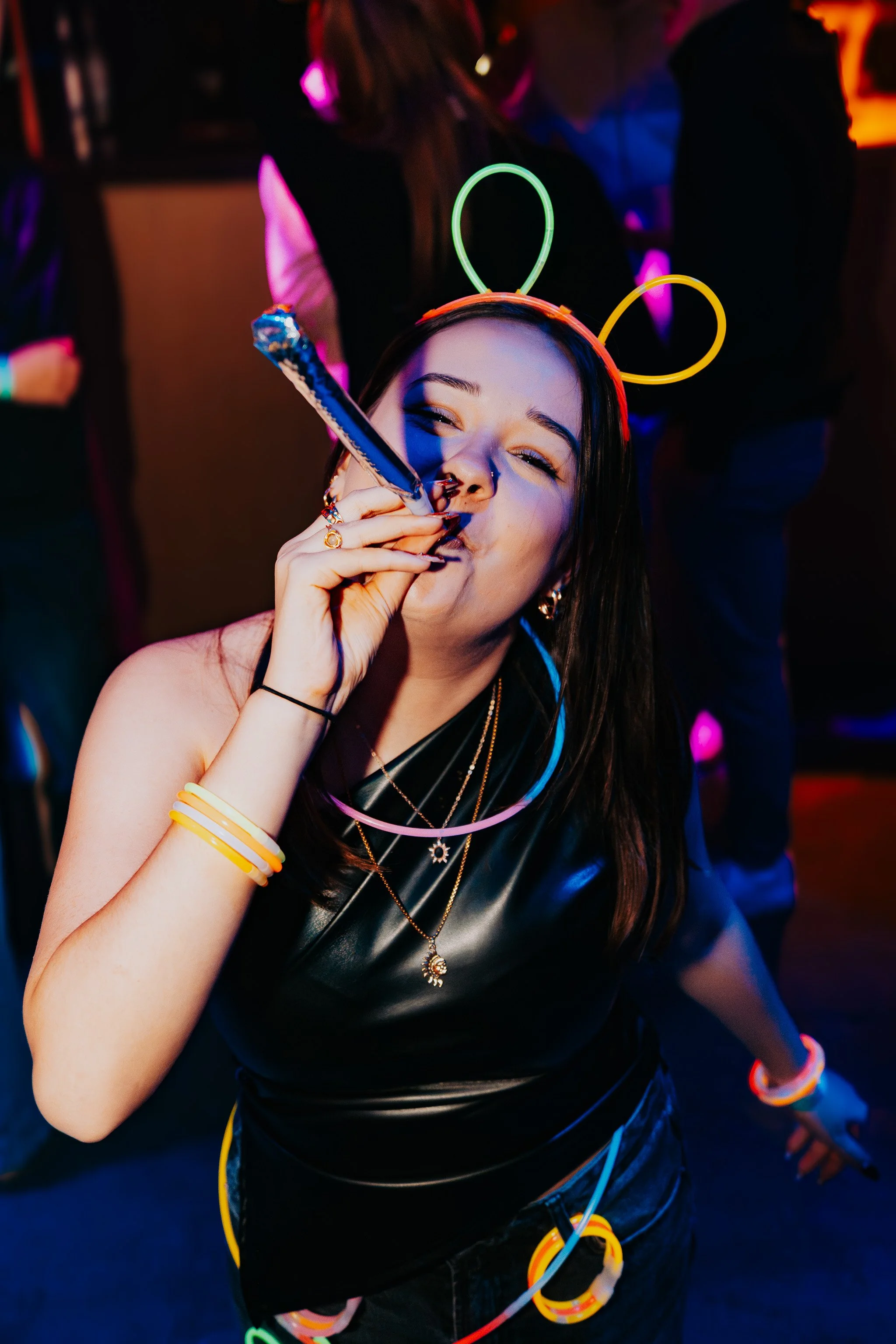 A woman with long dark hair wearing a neon light headband and colorful glow accessories dancing at a party under colorful lights.