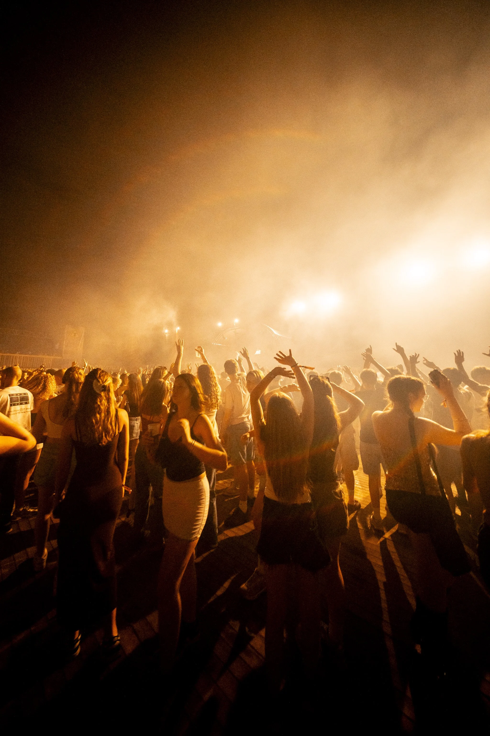 A large crowd of people at a concert or festival, facing a stage with bright orange and yellow lighting, raising their hands, with some visible painted faces and casual summer clothing.