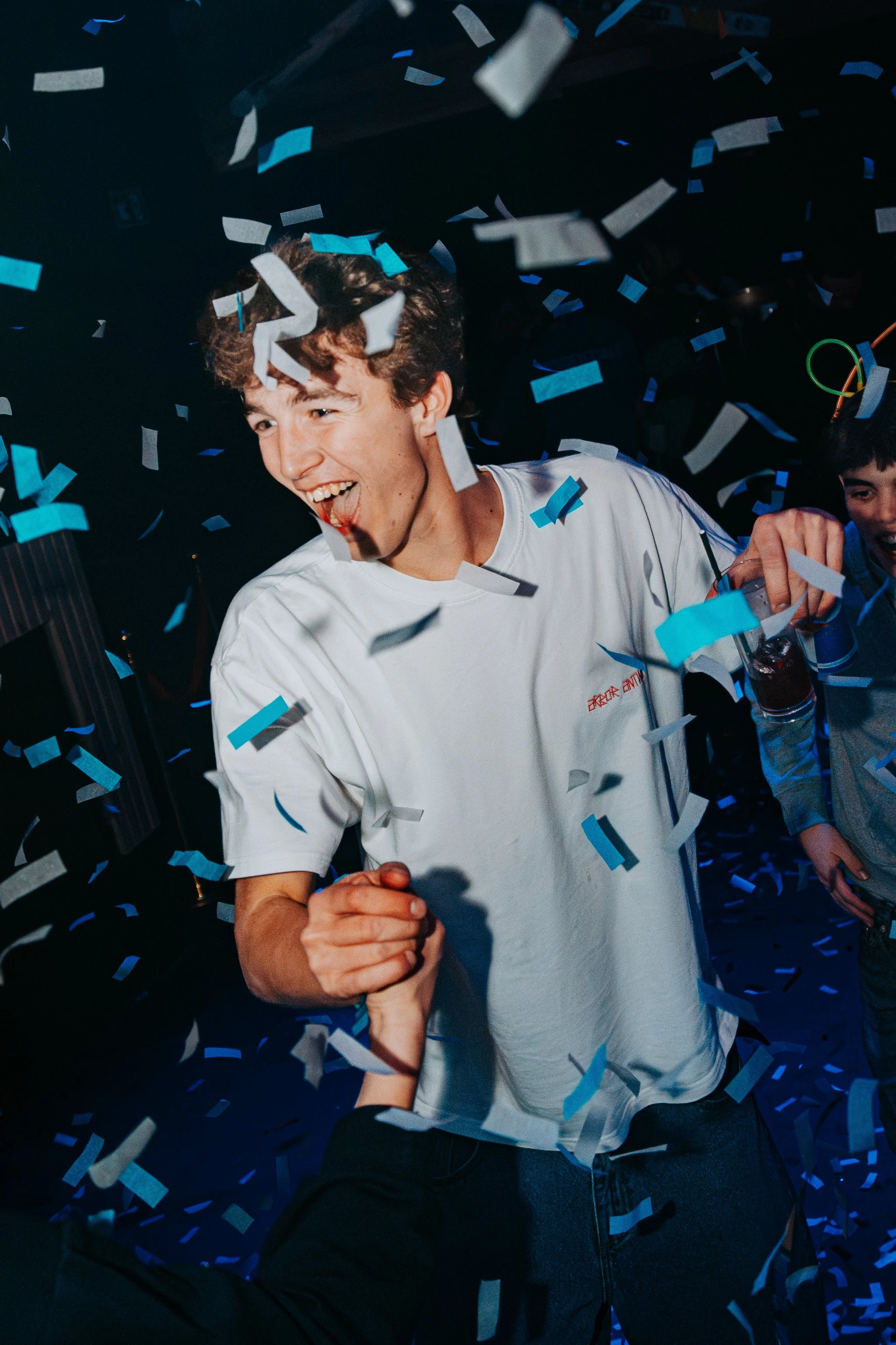 A young man with curly hair celebrating among falling blue and white confetti. He is smiling enthusiastically, wearing a white t-shirt, and holding hands with another person. The background is dark, suggesting a party or celebration environment.