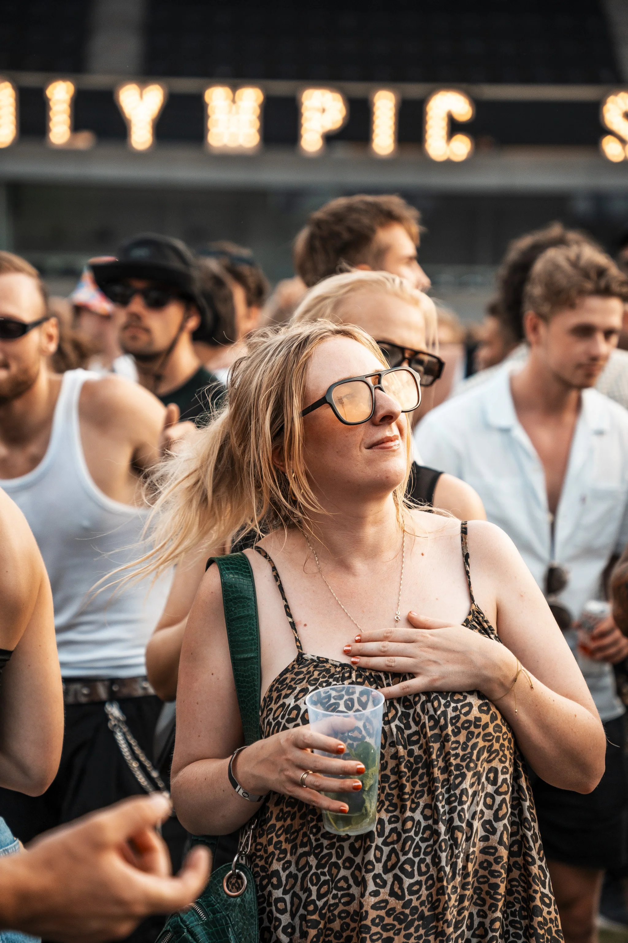 A group of people attending an outdoor event, with a woman in the foreground wearing sunglasses and a leopard print dress, holding a drink in her hand and placing her hand on her chest, with a crowd of people and festive lights in the background.