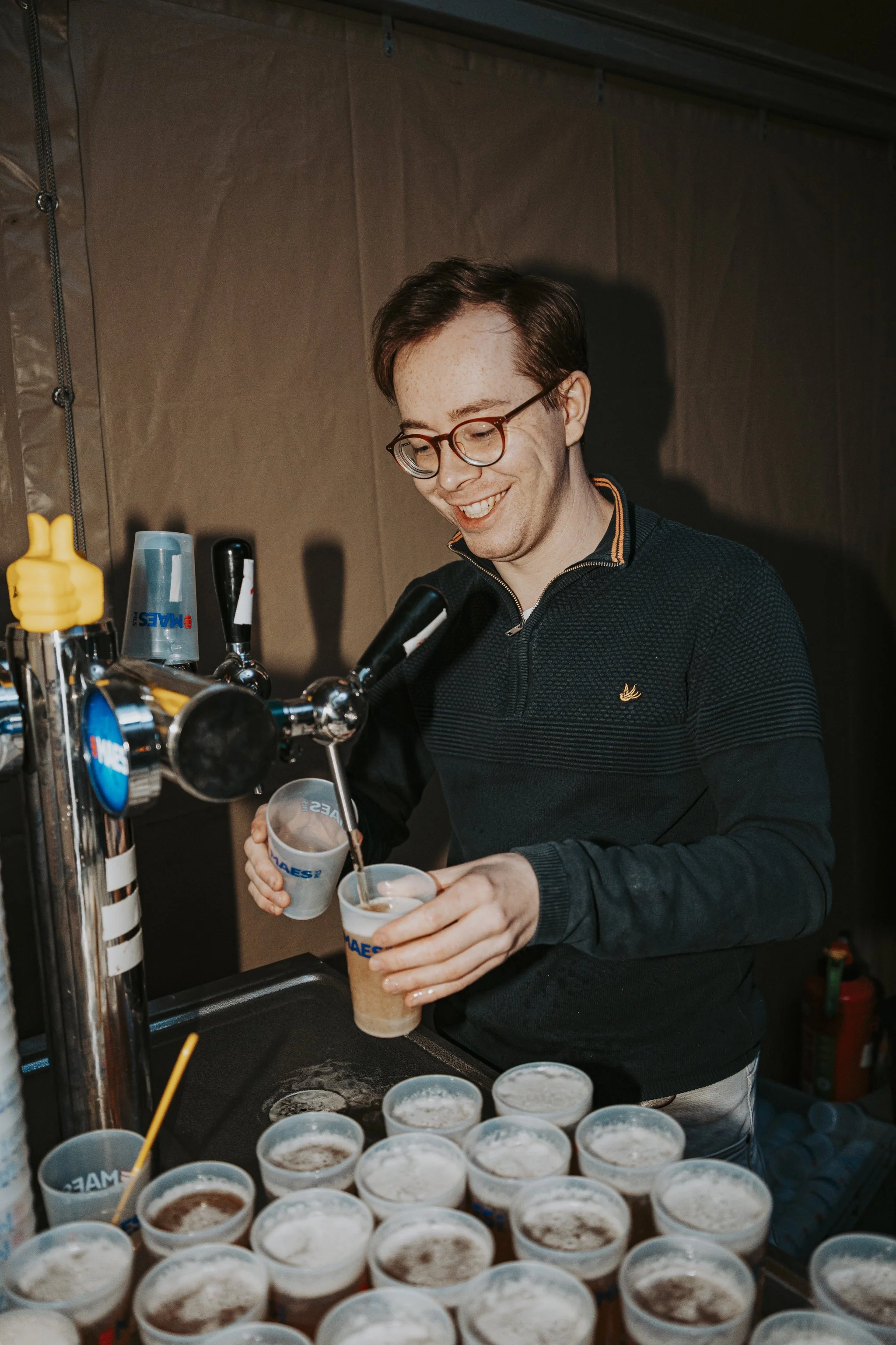 A young man with glasses and short brown hair is pouring a beverage into a clear plastic cup at a bar or beverage station. Various cups filled with drinks are arranged on the counter in front of him. The background shows a brown wall and part of a me