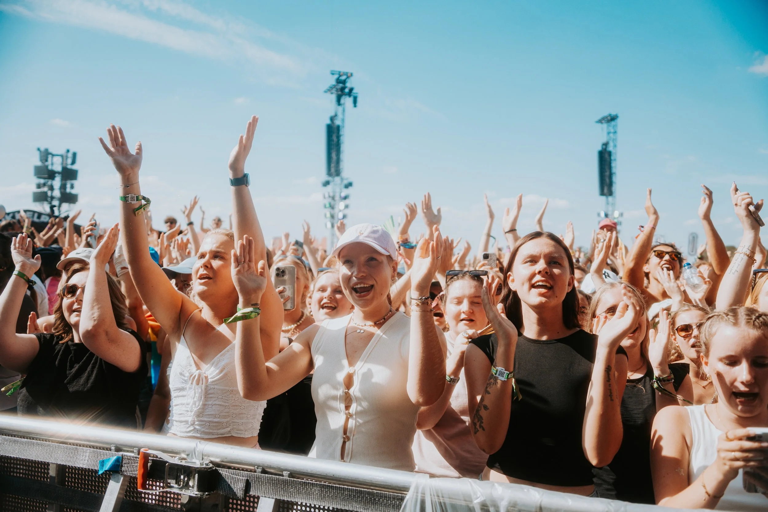 Crowd of young women at a music festival enjoying the performance with arms raised, outdoors on a bright sunny day.