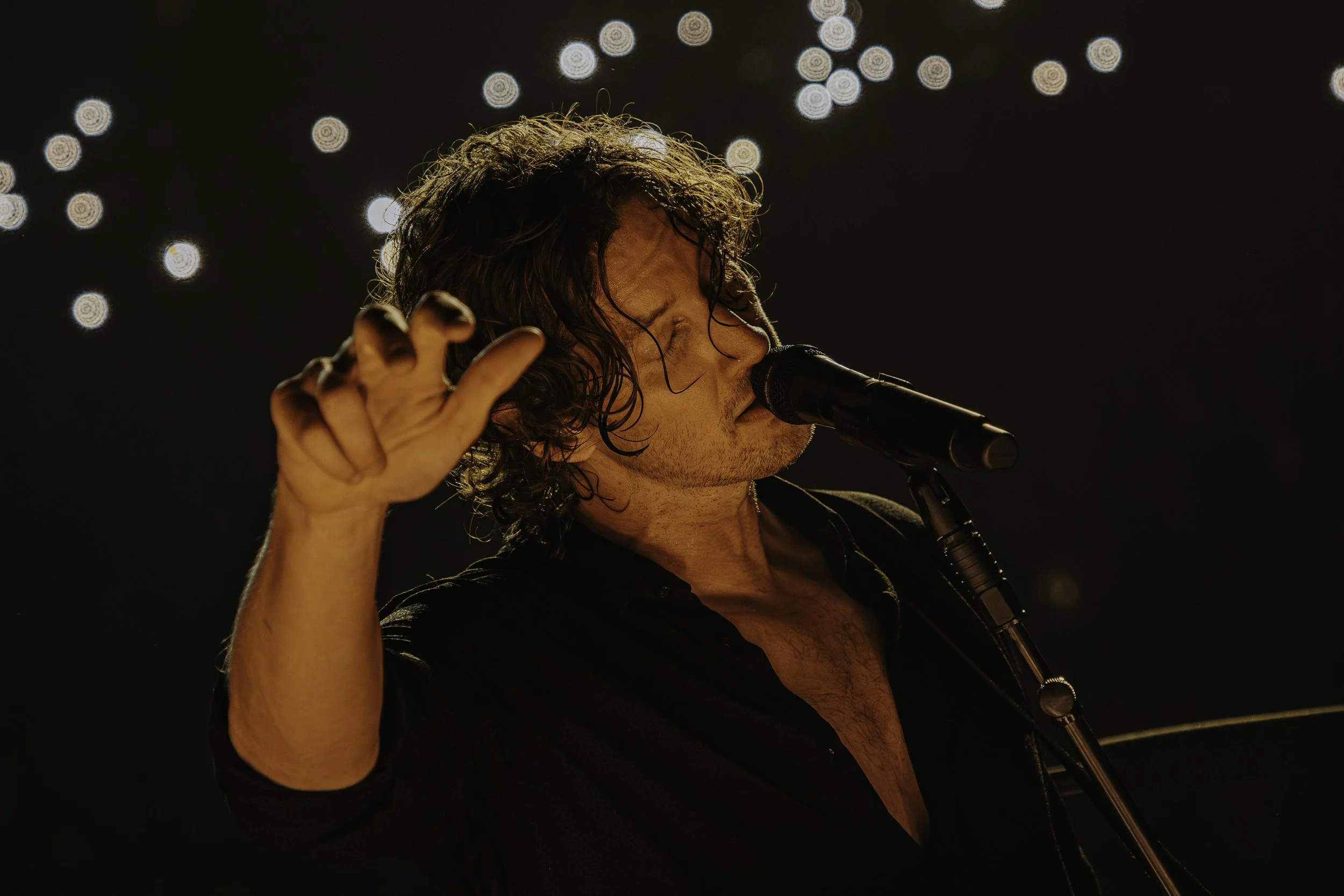 A male singer with wet hair passionately singing into a microphone on stage, with out-of-focus stage lights in the background.