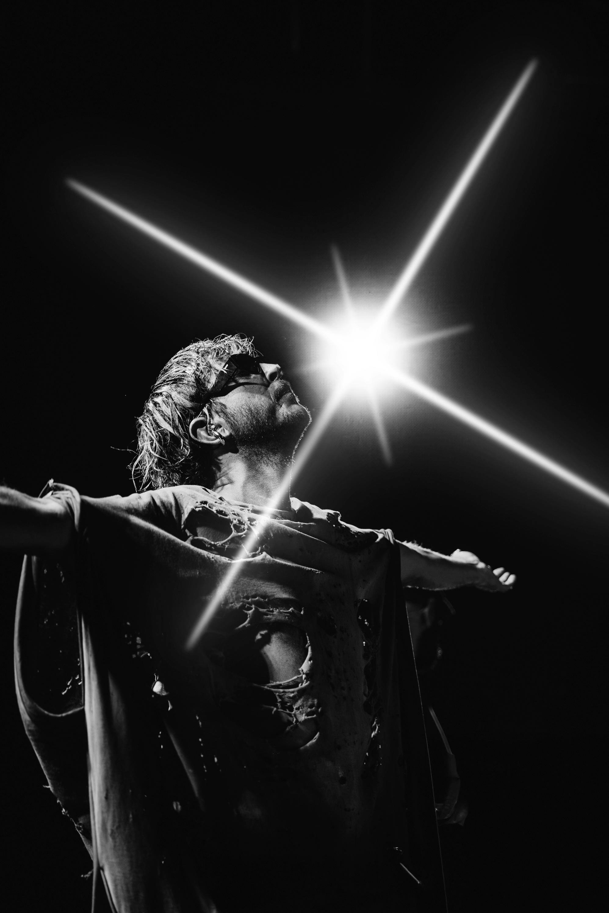 A person with glasses and wet hair, likely Jesus Christ, with arms outstretched, looking upwards, illuminated by bright light.