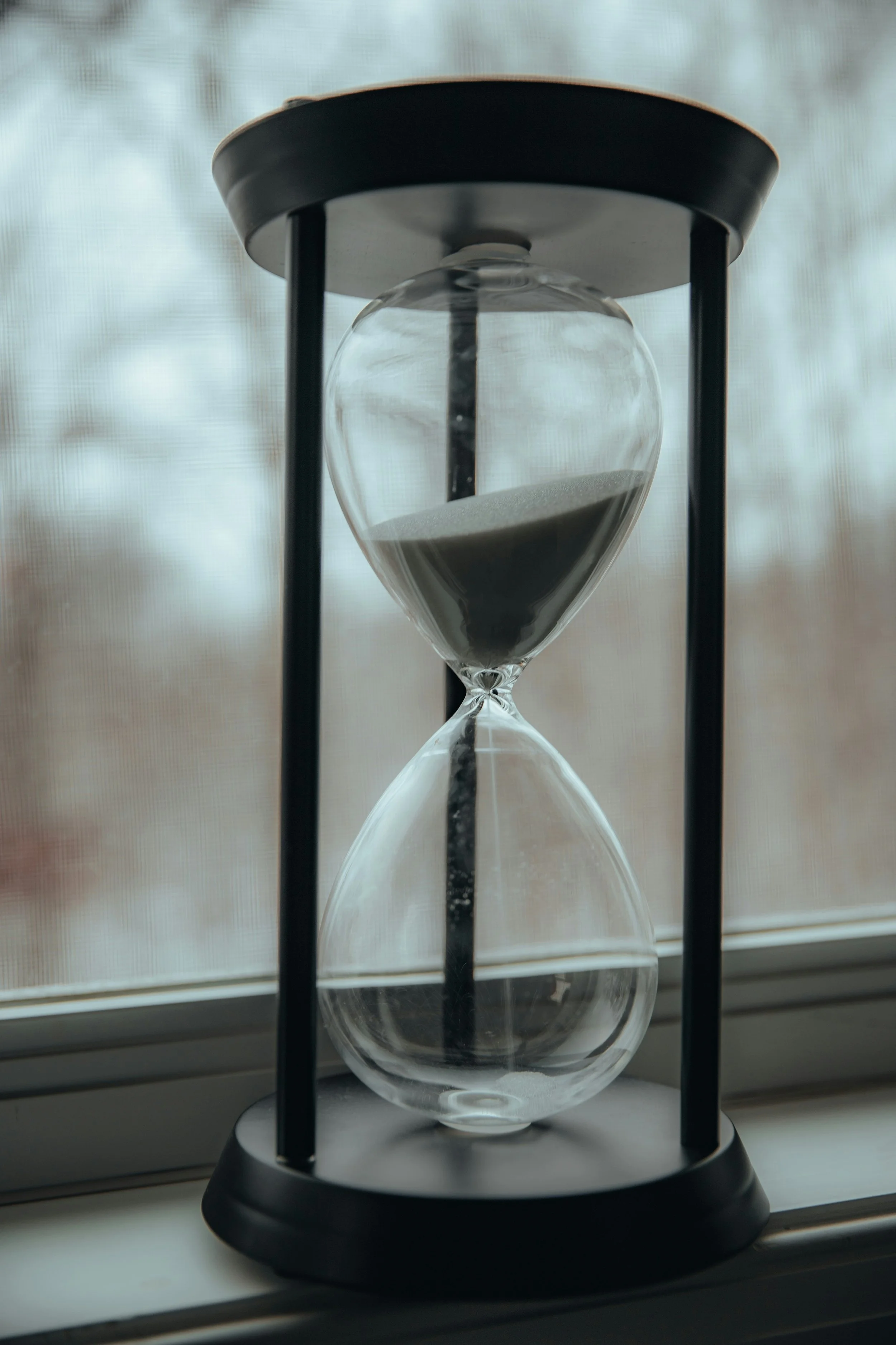 An hourglass with black frame and clear glass, filled with black sand, placed on a windowsill.