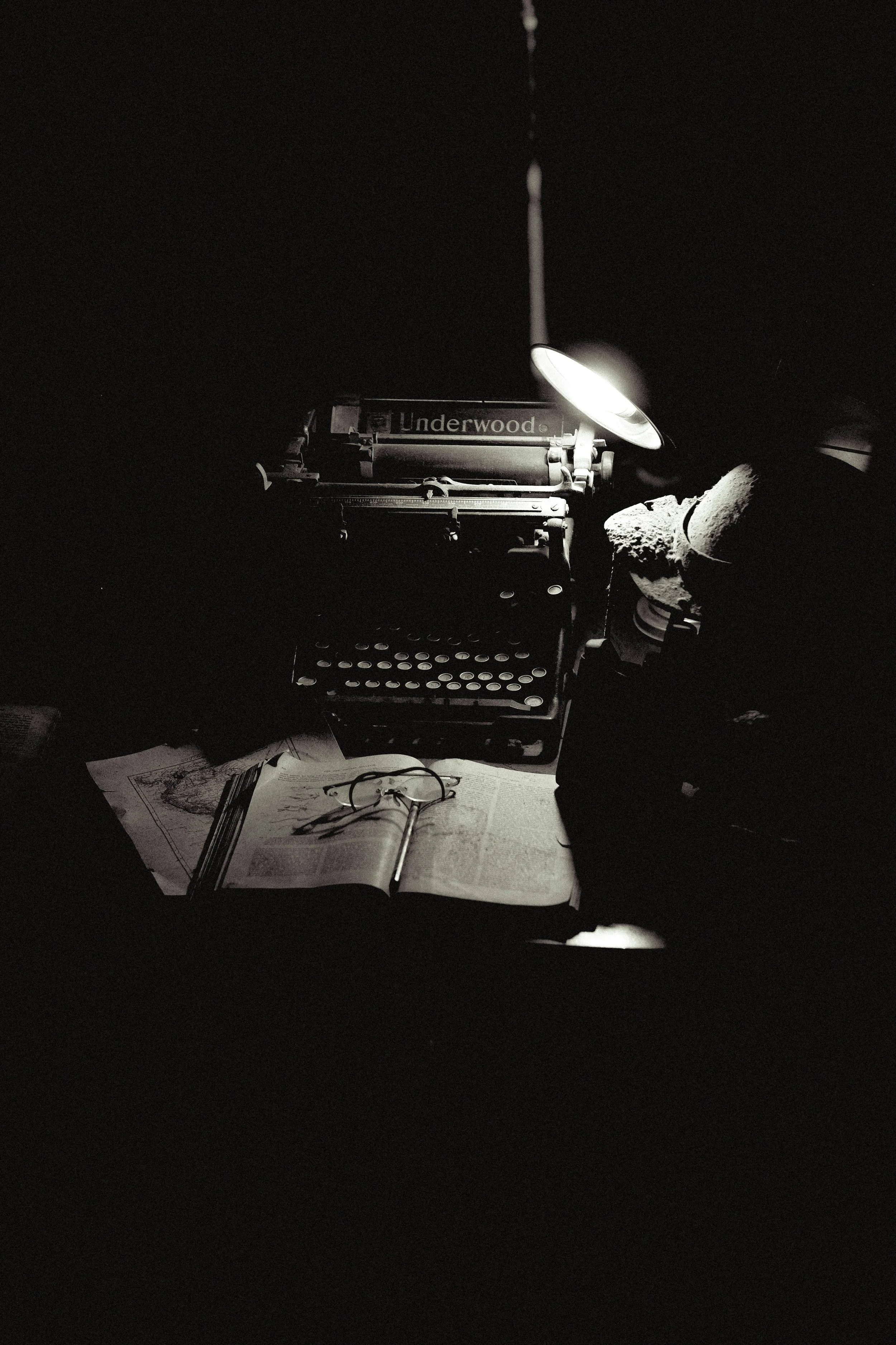 A vintage Underwood typewriter illuminated by a desk lamp, with glasses resting on an open book and papers underneath, in a dark setting.