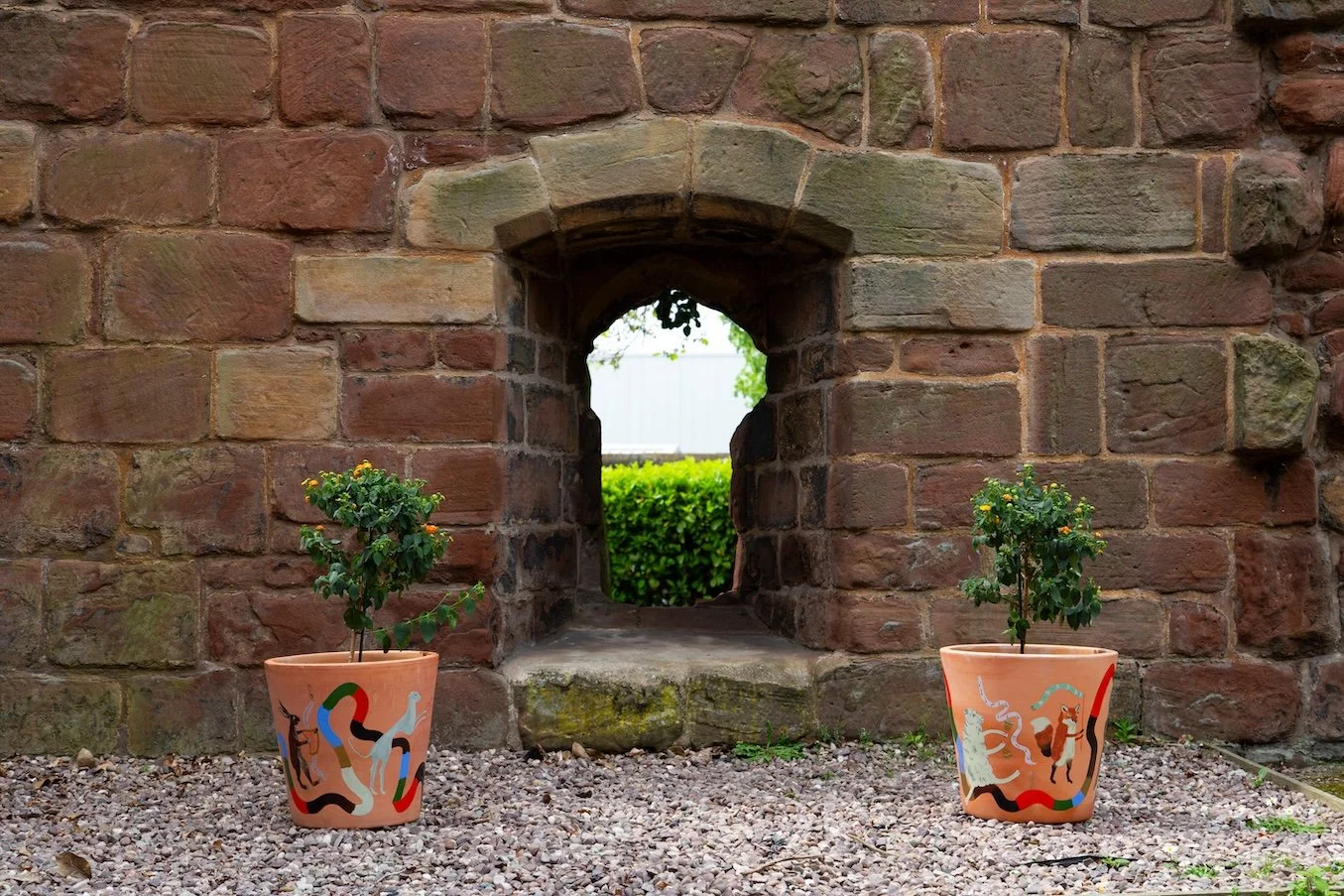 'ALMS WINDOW' Two large plant pots are situated in front of the ‘Alms Window’ as a welcome to the visitors.  The pots depict a procession of animals and people inspired by medieval iconography.  A parallel is drawn between the pilgrimage to the Prior