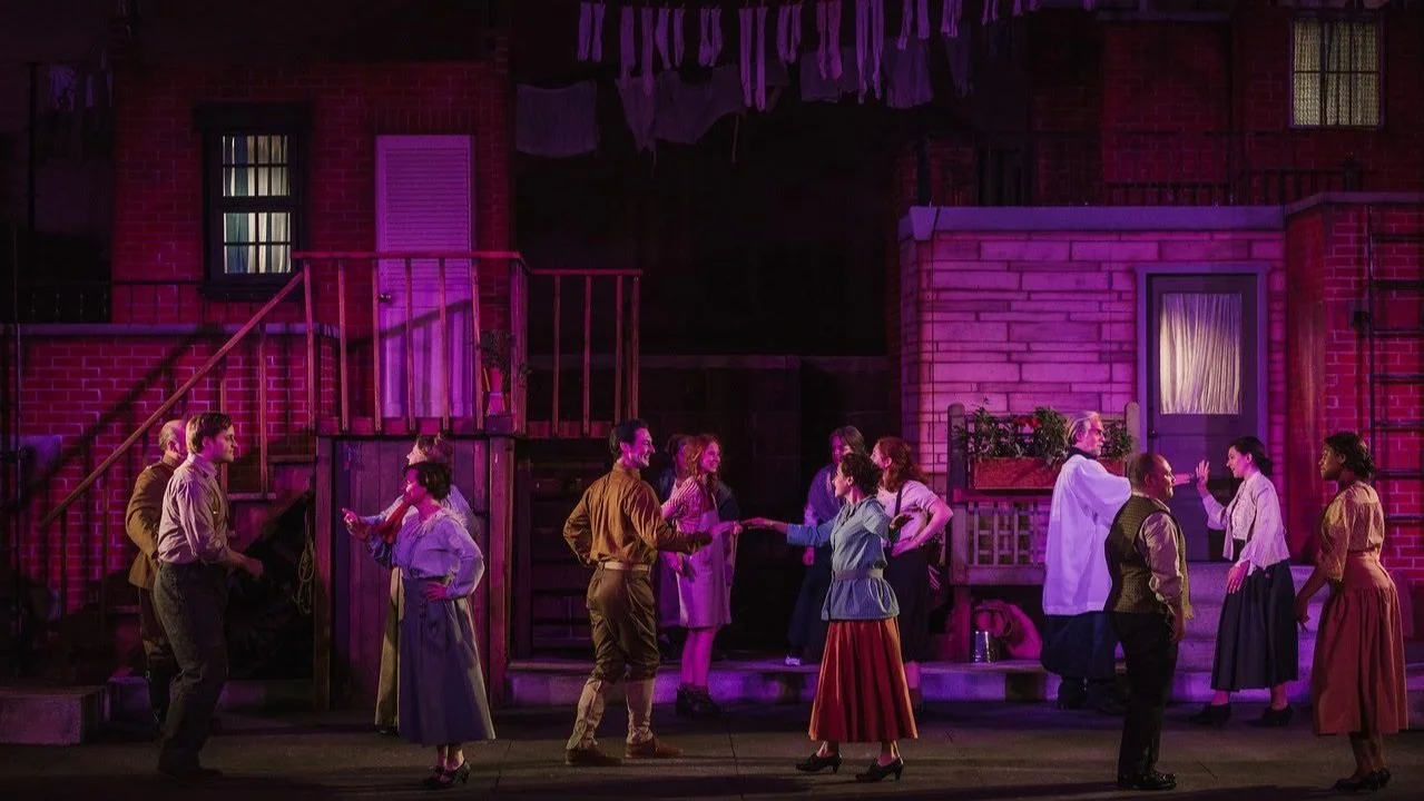 Stage scene of a theatrical production depicting a street with actors in period costumes engaging in various interactions, with a backdrop resembling apartment buildings.