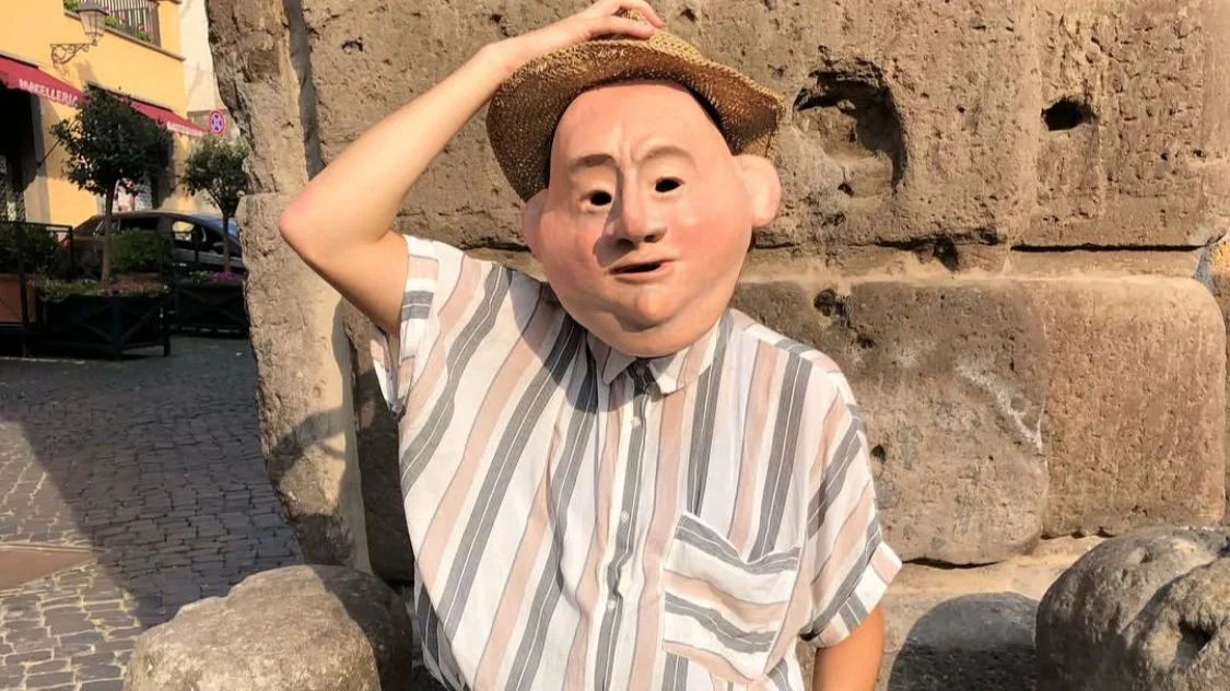 A person wearing a strange, exaggerated mask with a large face, small eyes, and a wide mouth, holding a straw hat with one hand, standing outdoors next to a stone wall.