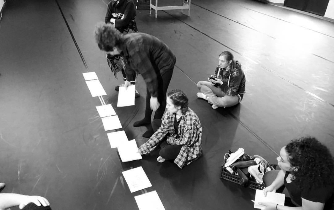 Group of young women sitting or kneeling on the floor, engaging in an activity involving papers laid out in a line, in a room with a dark floor and minimal furniture.