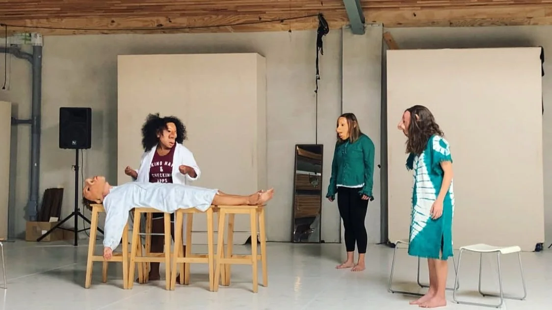 Three women in a studio with a wooden ceiling, two of them are watching as the third woman and another person on a table lie still. The woman on the left, with curly hair, appears to be demonstrating or explaining something. The person on the table is covered with a white cloth, and they seem to be part of a performance or theatrical scene.