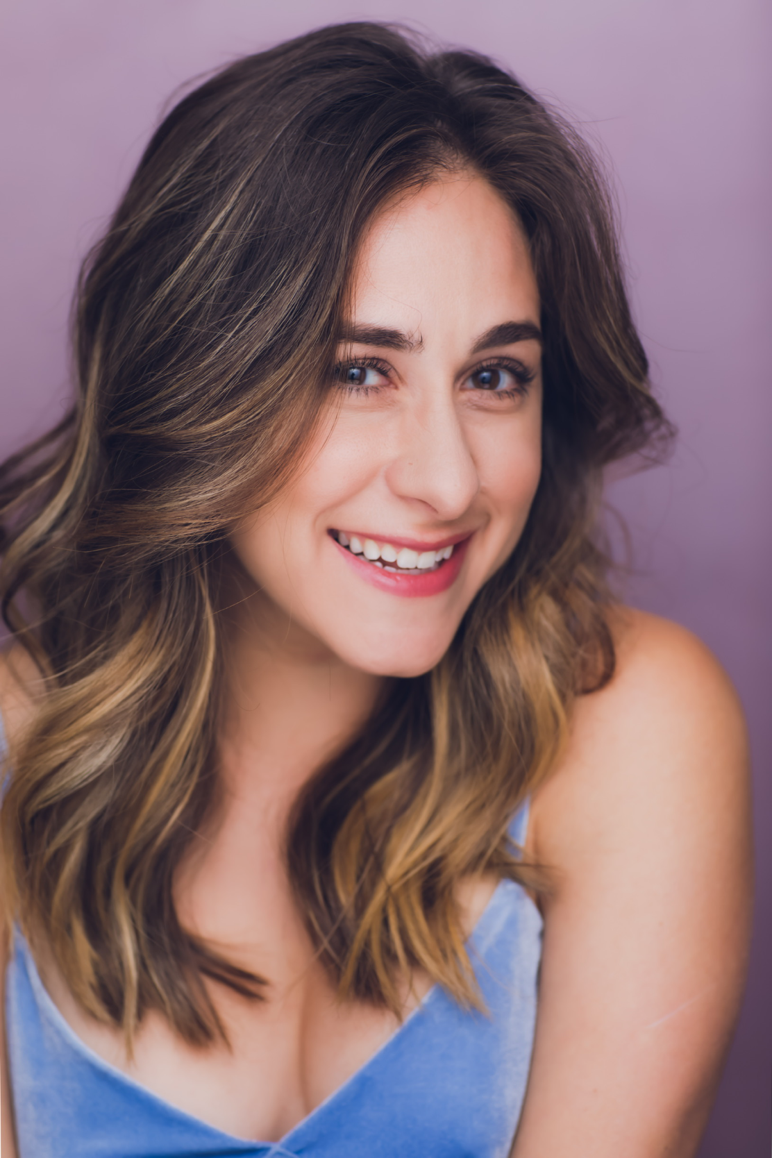 A young woman with shoulder-length wavy brown hair, smiling, wearing a light blue top, against a purple background.