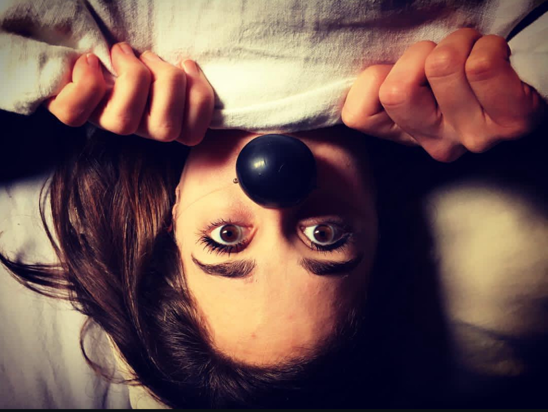 A woman lying upside down, wearing a white shirt, with a black nose clip on her nose, holding the shirt with both hands.