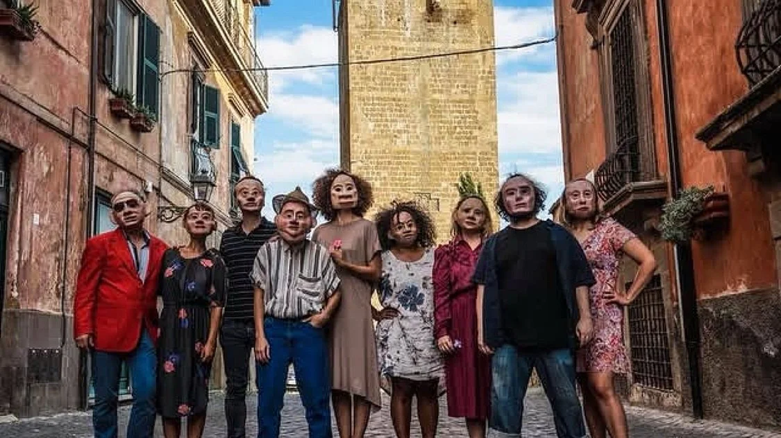 A group of nine people wearing masks with exaggerated facial features, standing on a cobblestone street in an alleyway with old buildings and a tower in the background.