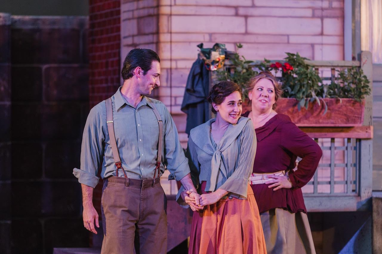 Three actors performing on stage, with two women and one man, in a scene with a brick wall and flower box in the background.