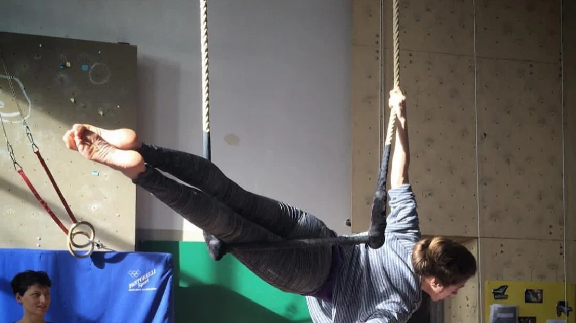 Young woman practicing aerial trapeze routines in a gym, hanging upside down with her legs wrapped around the fabric, and a wall with climbing holds in the background.
