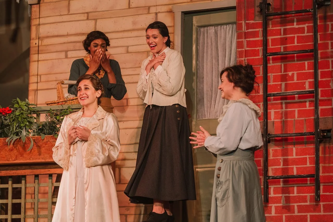 Four women in vintage clothing celebrating on a porch, with one woman smiling, one covering her mouth in surprise, and another clapping.