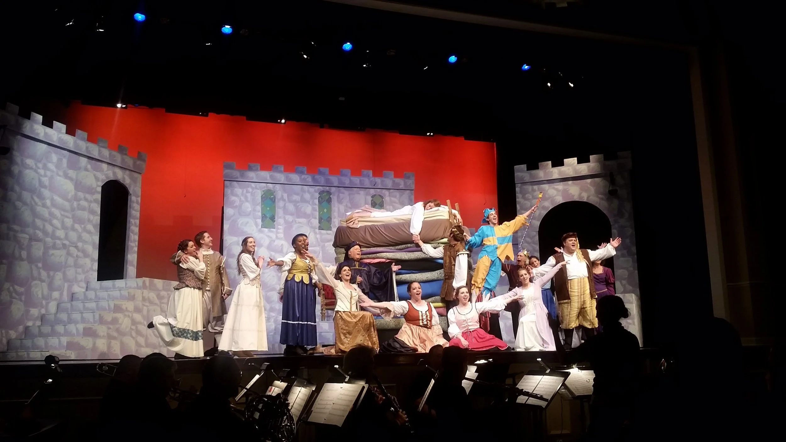 A stage performance featuring actors dressed in medieval-style costumes, with castle-themed backdrop. One actor is dressed as a jester, while others are singing or acting with expressive gestures. Musicians are silhouetted in the foreground.