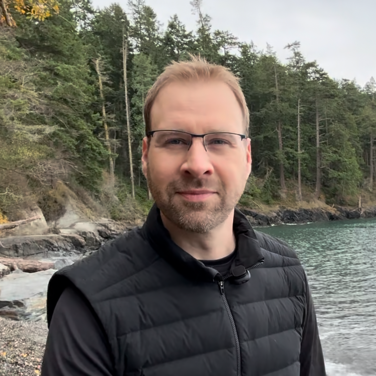 A man with glasses and a black jacket standing outdoors near a body of water with a forested shoreline in the background.