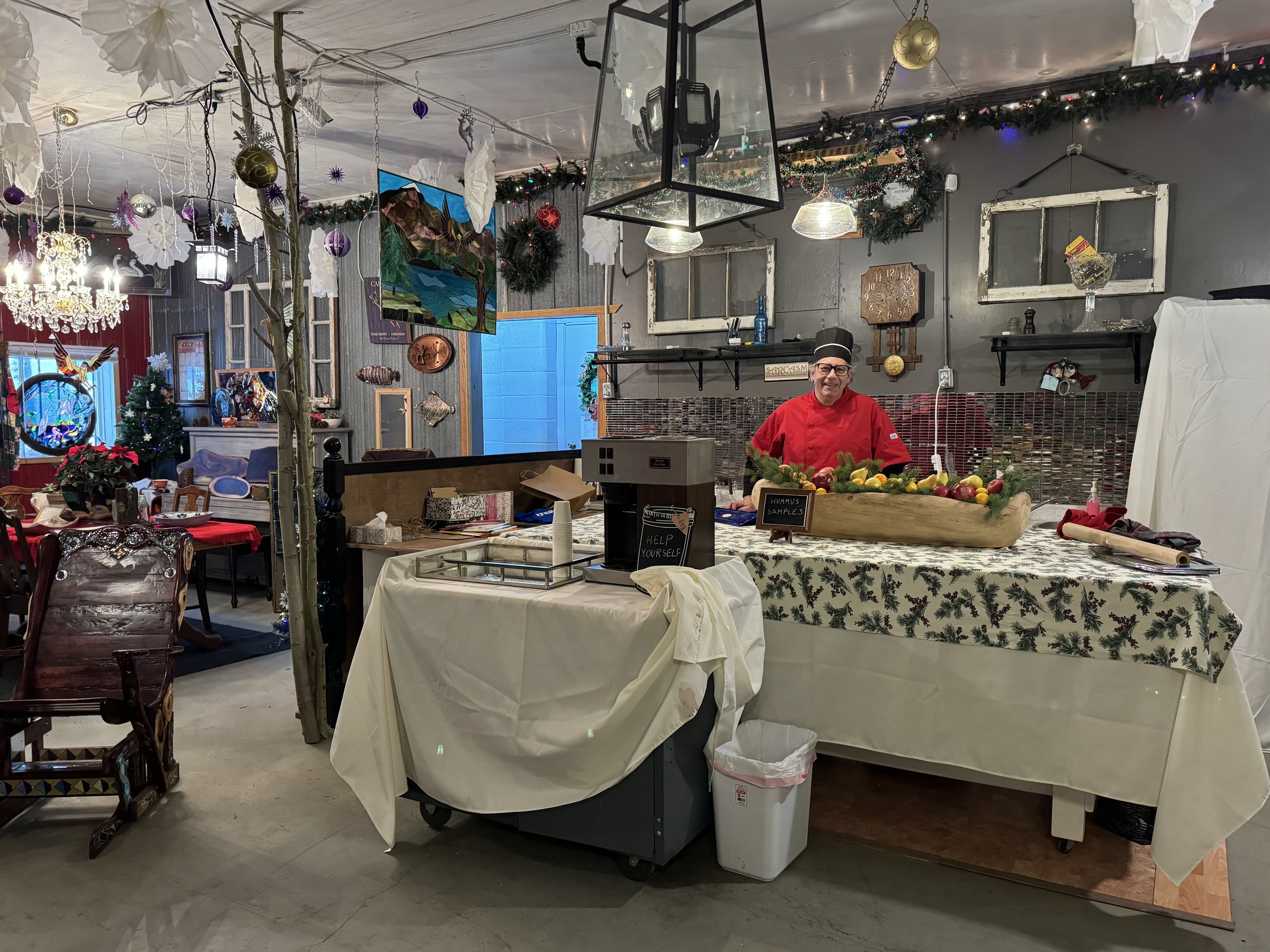A person in a red chef's coat standing behind a table with a festive holiday tablecloth, displaying a variety of fresh vegetables and herbs. The setting is decorated for Christmas with ornaments, wreaths, and holiday lights inside a cozy, decorated r