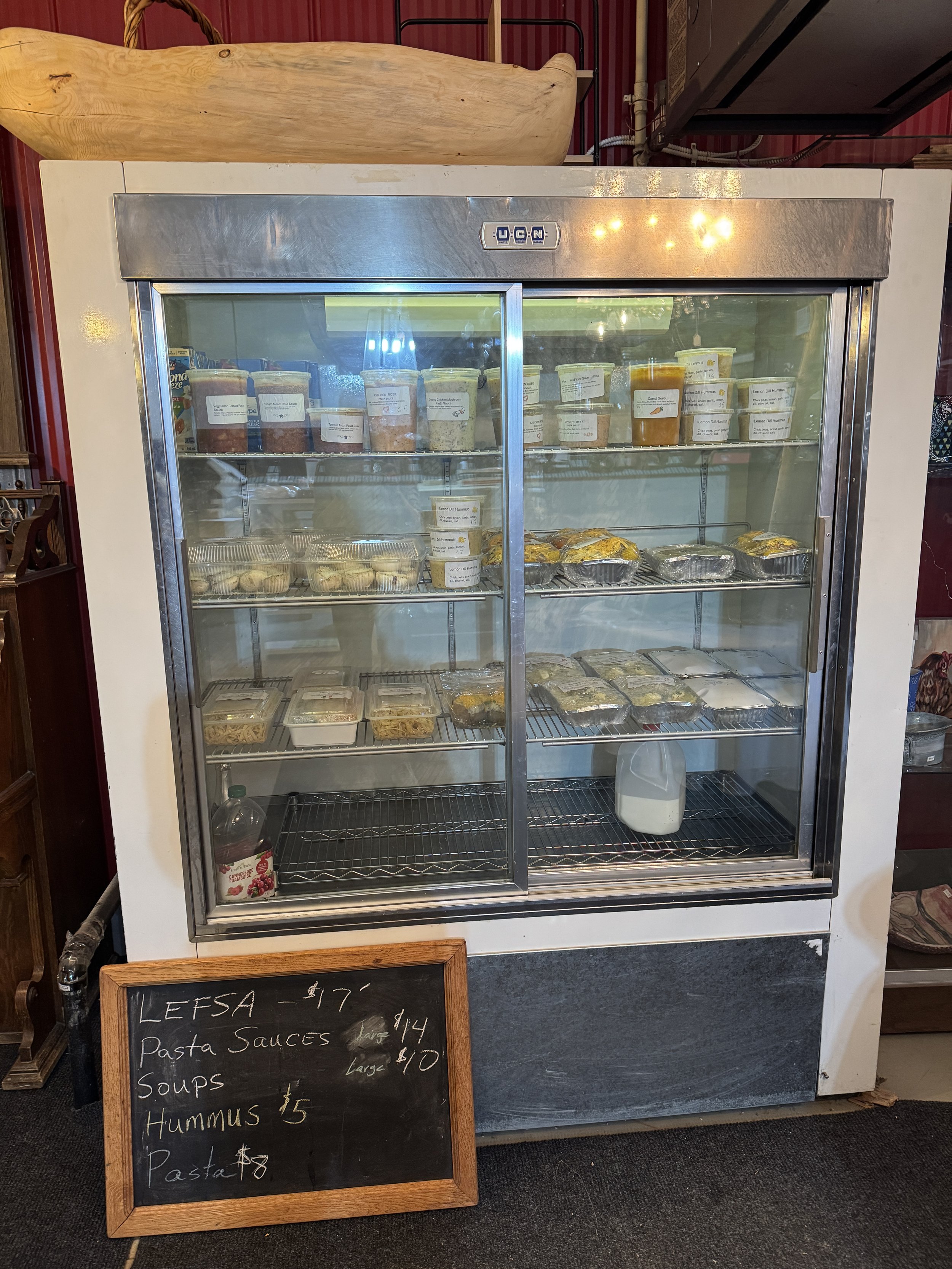 Partially filled refrigerated display case with pre-packaged food items, including pasta and soup containers, and a small chalkboard sign listing food items and prices in front.