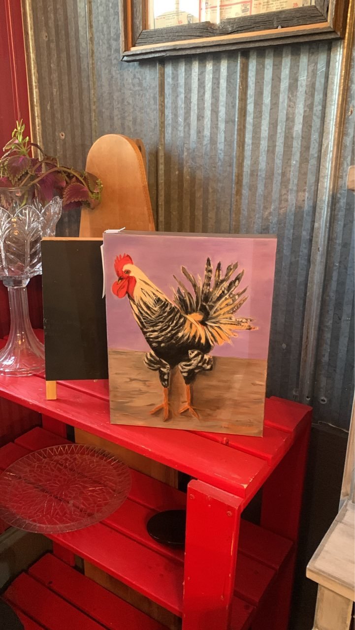 Painting of a rooster with colorful feathers, set against a pink background, displayed on a red shelf in a rustic setting.