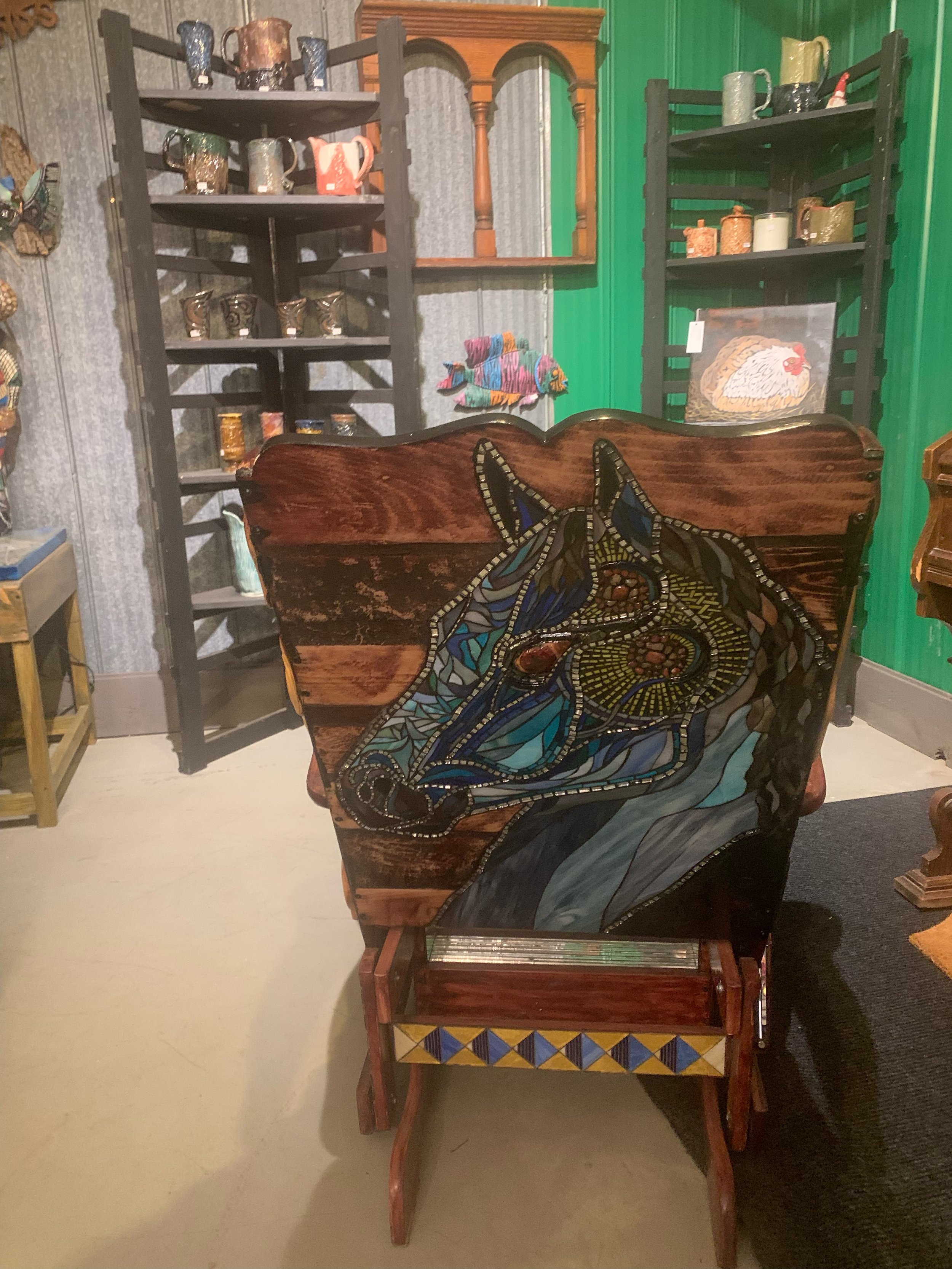 A wooden chair with a mosaic of a horse's head painted on the backrest, surrounded by shelves filled with pottery and decorative items in a rustic room.