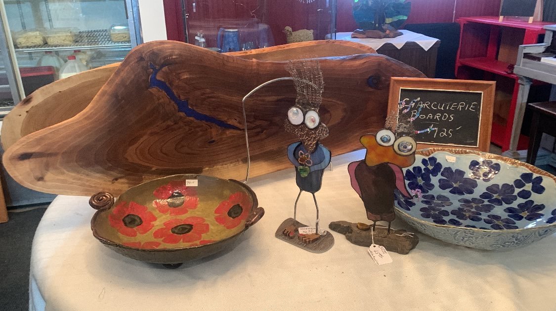 Decorative wooden and ceramic art pieces, including a large wooden board, a ceramic bowl with red poppy flowers, two abstract metal figurines, and a ceramic bowl with blue floral patterns, displayed on a table in an artisan shop.