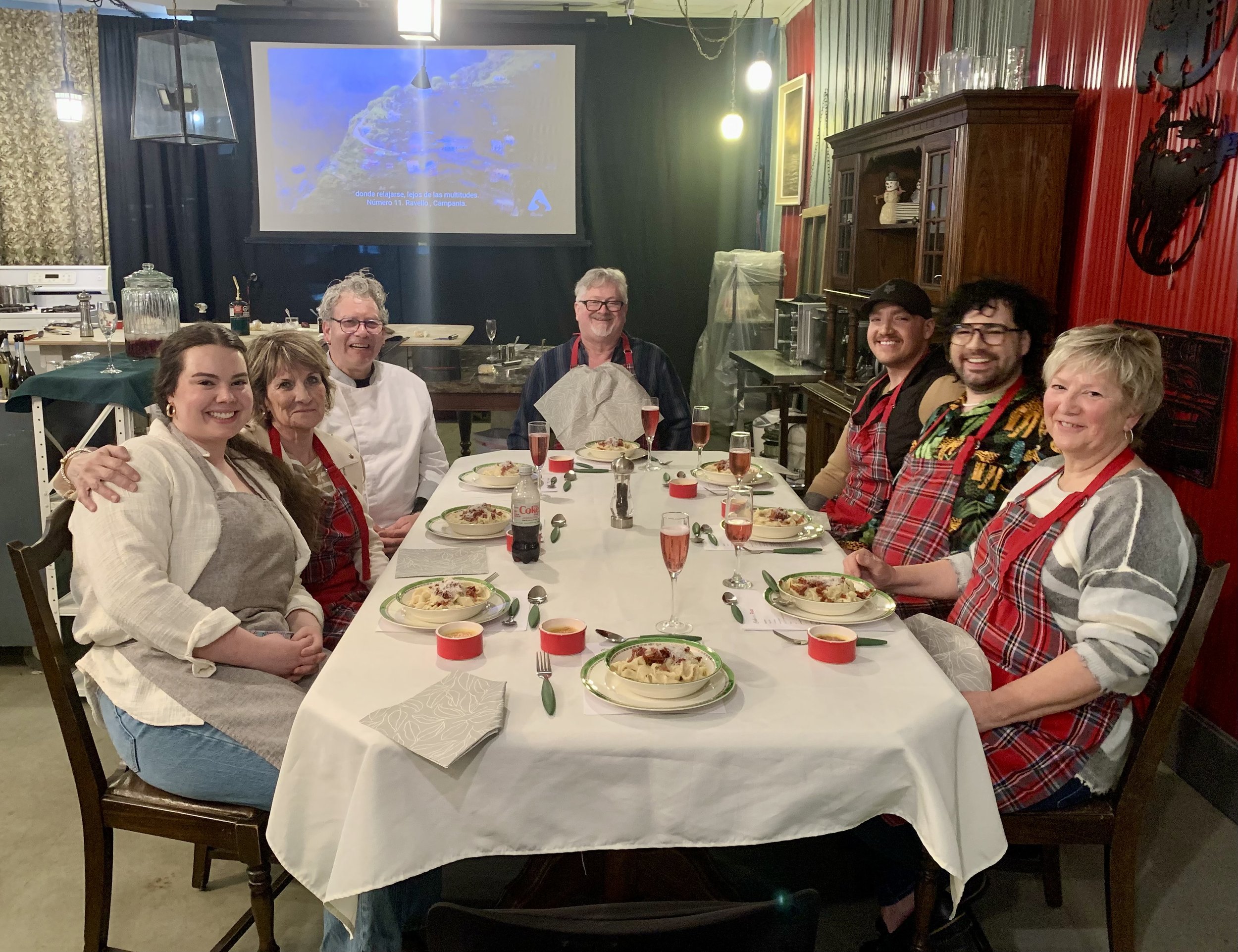 A group of nine people sitting around a dinner table in a cozy restaurant, smiling at the camera. The table is set with plates of pasta, glasses of pink drinks, and red candles. The restaurant decor includes red walls, wooden furniture, and a large s