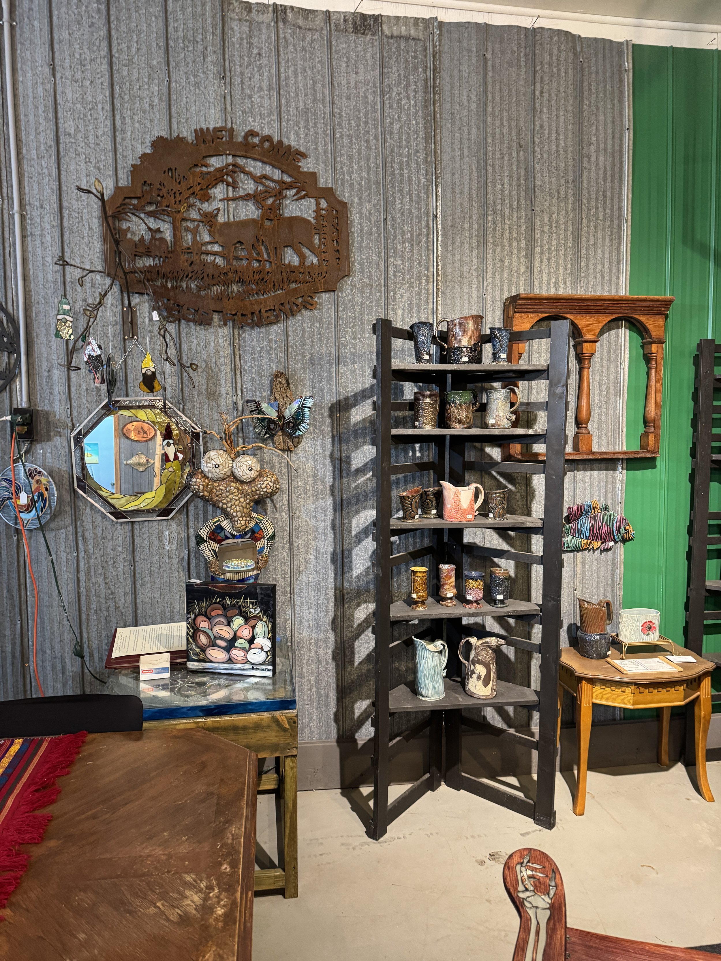 Indoor display of various crafts and decorative items, including pottery, wall art, and jewelry, on shelves and tables against a metal wall with a Welcome deer sign.