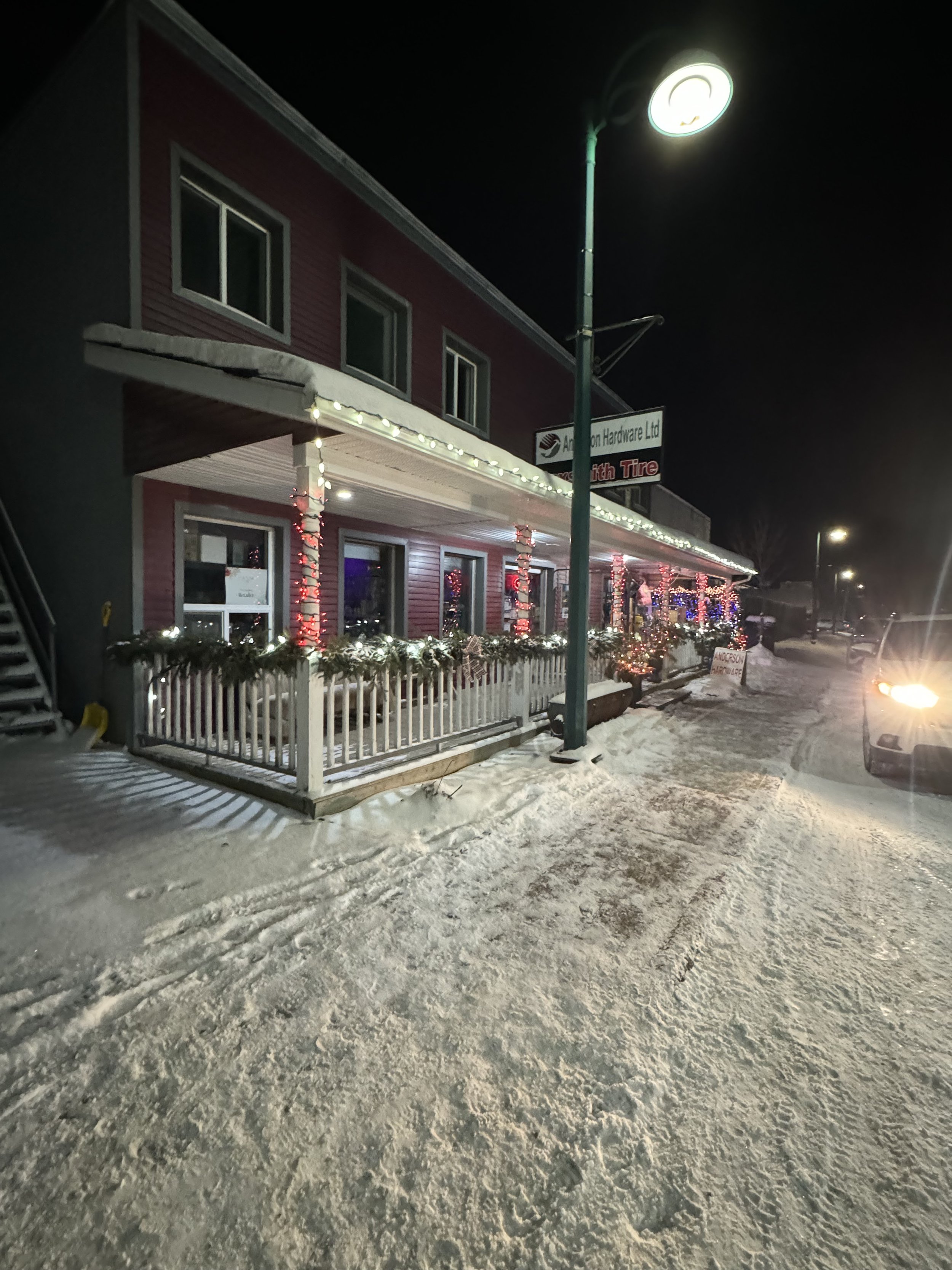 A snow-covered sidewalk and street at night with a building decorated with Christmas lights and garlands, and a streetlamp illuminating the area.