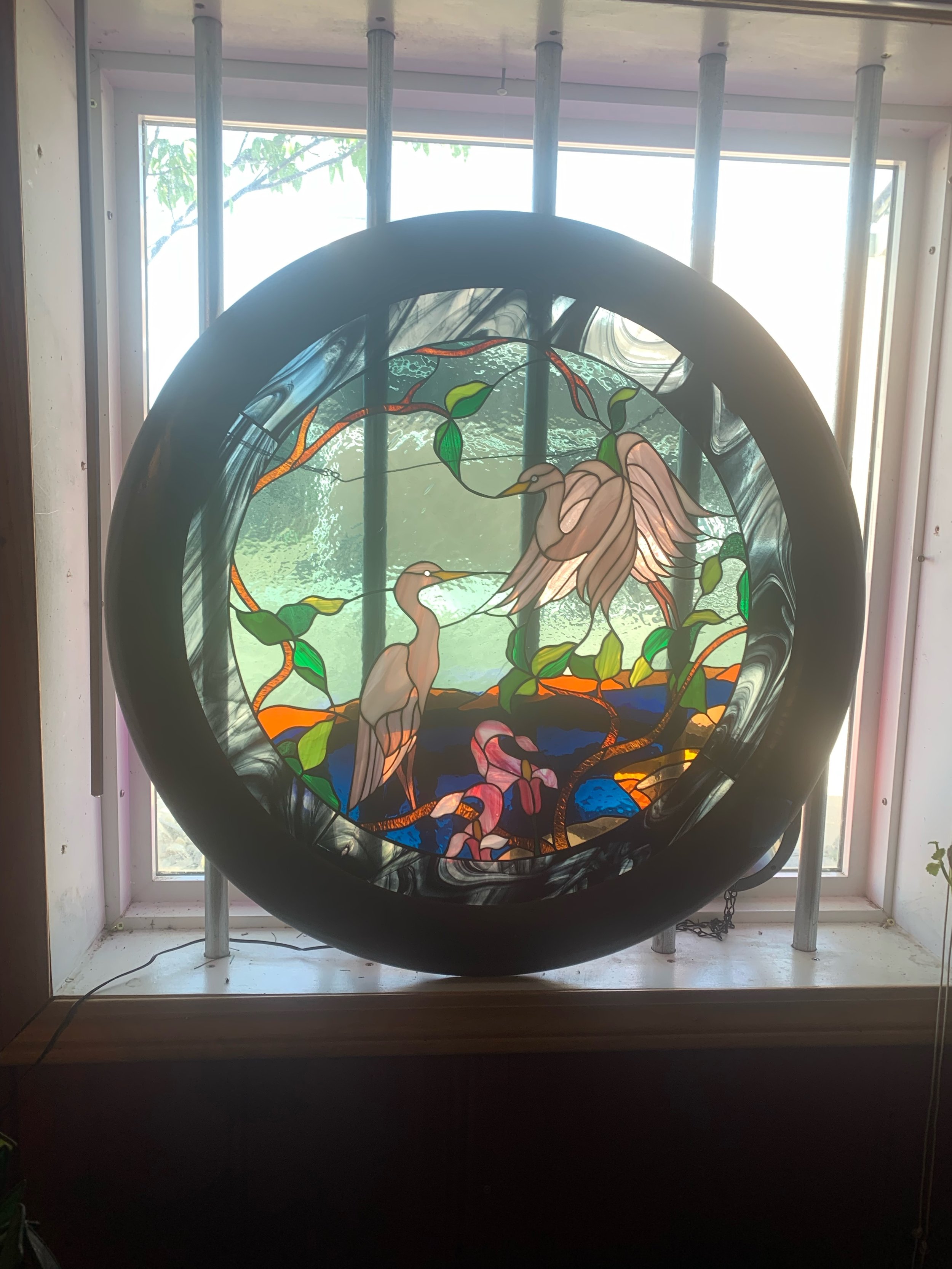 Stained glass window depicting two herons standing among trees and colorful plants.