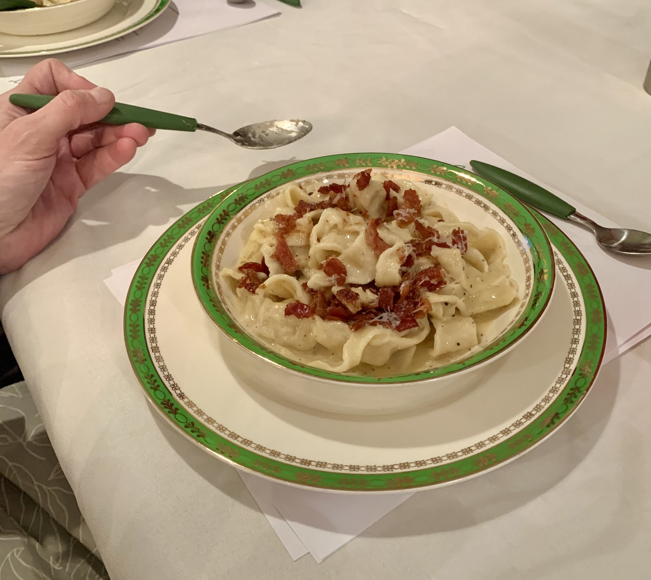 A bowl of cooked pasta with bacon bits on top, served on a decorative green and gold plate, on a white tablecloth with a hand holding a spoon.