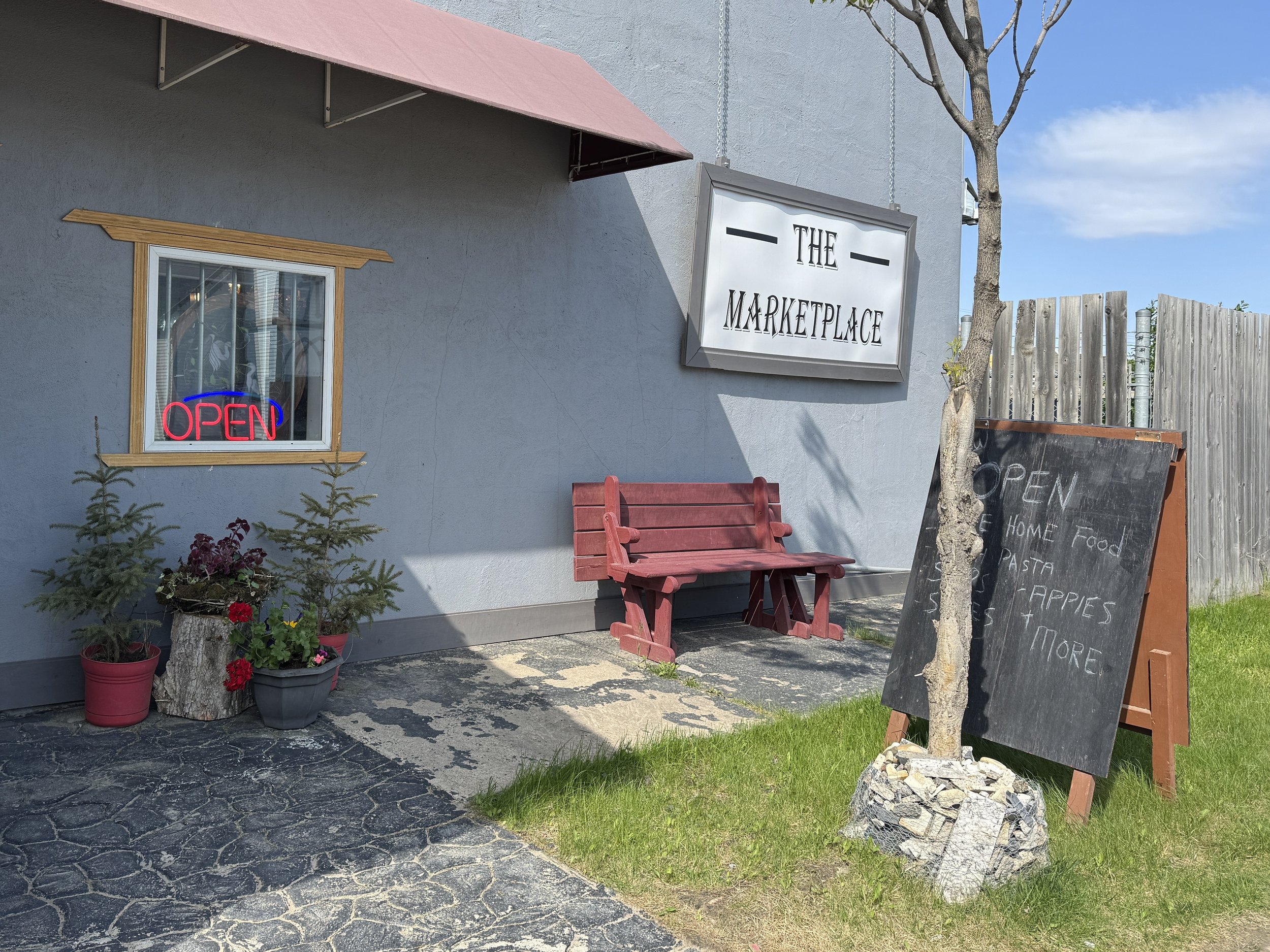 Exterior of a marketplace with a gray building, a window with a neon 'OPEN' sign, a red bench, potted plants, and a black chalkboard sign listing food items, next to a wooden fence and a small tree.