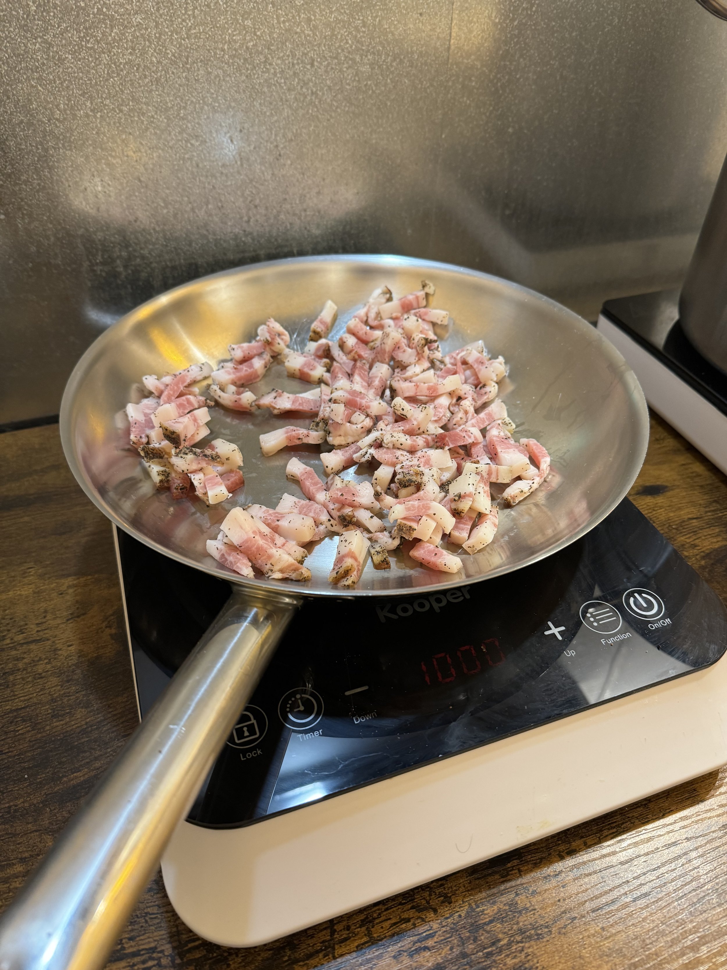 Cooking small chopped bacon pieces in a stainless steel pan on an electric stovetop.