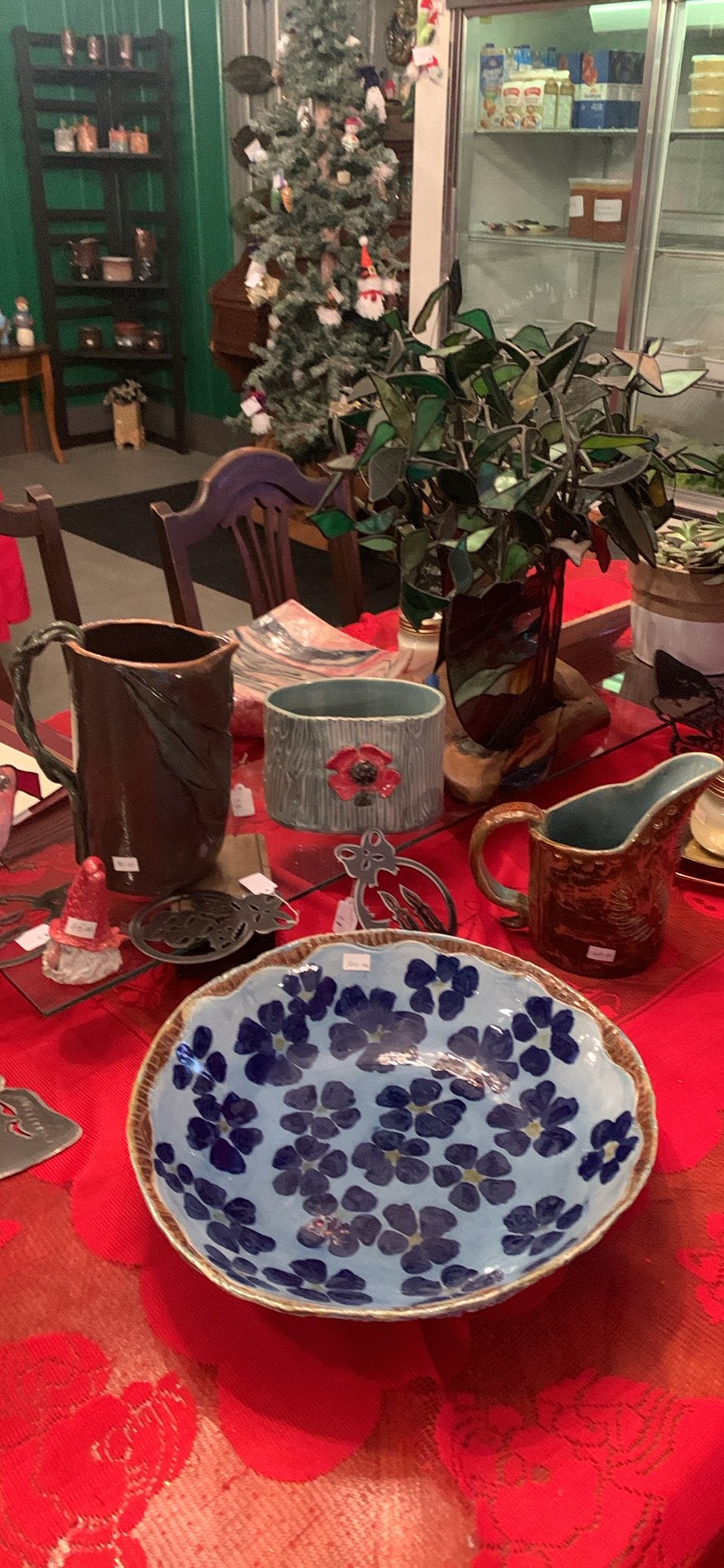 Display of Christmas-themed pottery and ceramics on a red tablecloth, with a decorated Christmas tree in the background and a refrigerator with various food items inside.