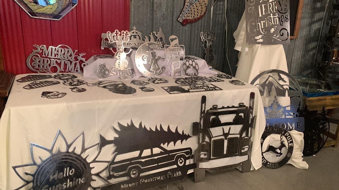 A table displaying holiday-themed metal signs and decorations, including a "Merry Christmas" sign, reindeer, paw print, and truck motifs, with some displayed on a white tablecloth and others in front of a red wall.