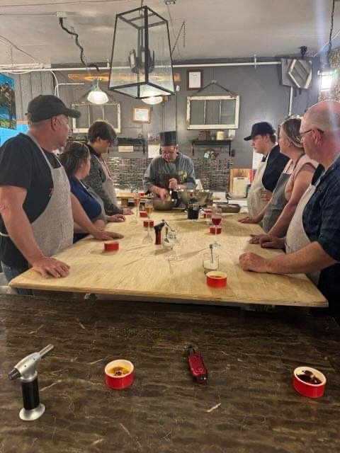 A group of people gathered around a large wooden table in a kitchen setting, participating in a cooking class led by a chef. The chef is demonstrating a cooking technique, with ingredients and utensils on the table, and participants closely watching.