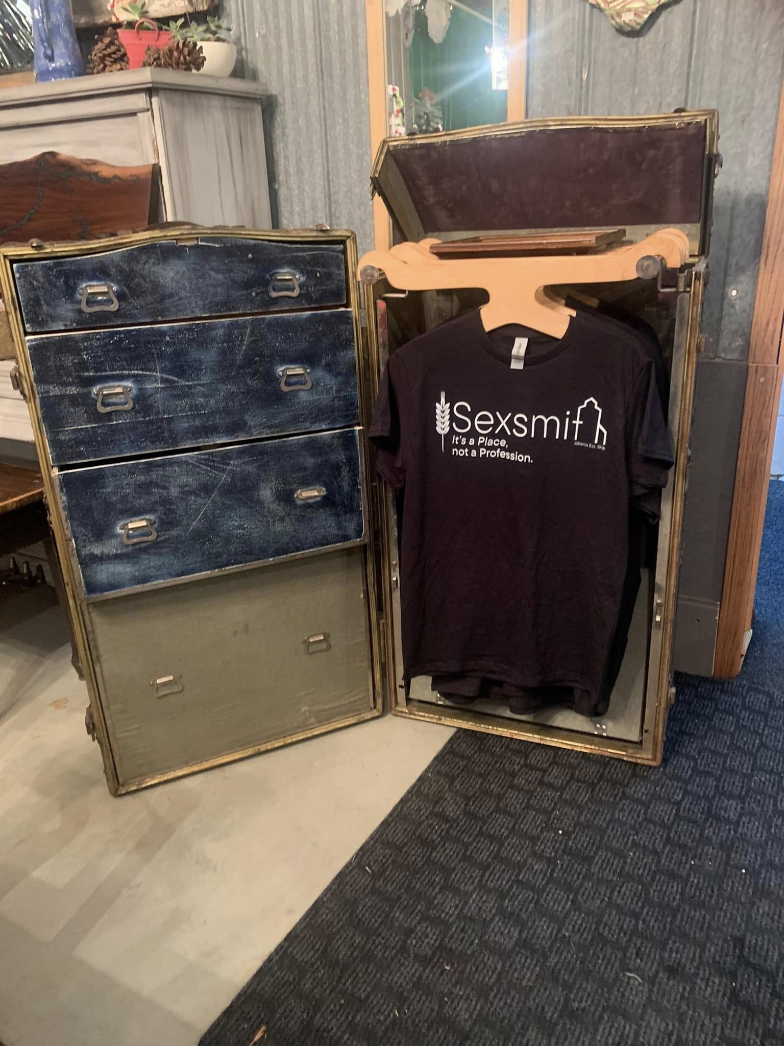 Open vintage-style metal locker with a black T-shirt inside that has text and an outline of a house, set in an indoor space with a carpeted and wood-paneled floor, and other furniture and decorations in the background.