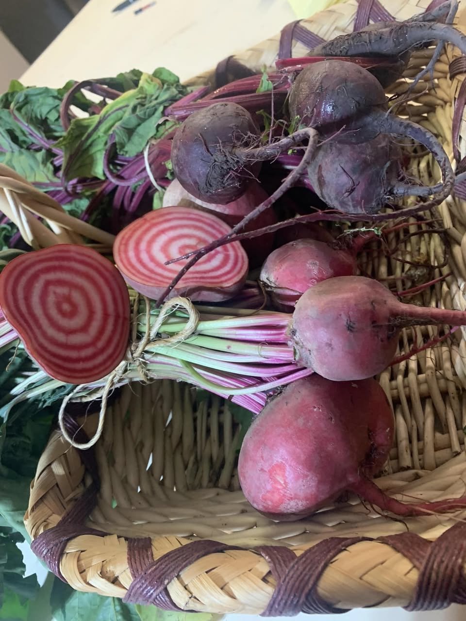 A basket filled with fresh beets, including whole beets with roots and leafy greens, and some sliced beets showcasing their red and white spiral pattern.