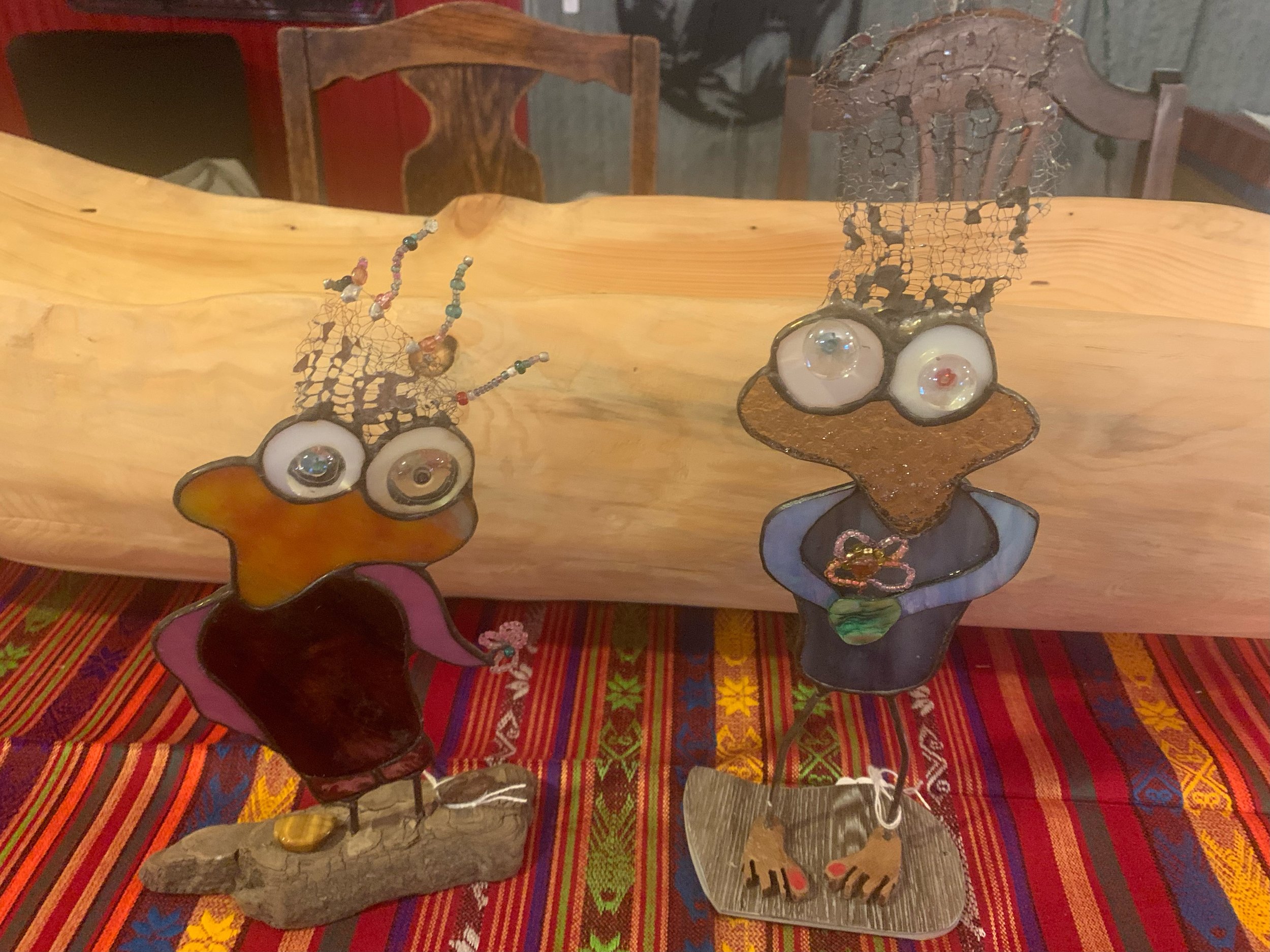 Two whimsical metal and glass wall art figurines of humorous cartoon characters with exaggerated eyes, placed on a colorful woven tablecloth with a wooden backdrop and chairs in the background.